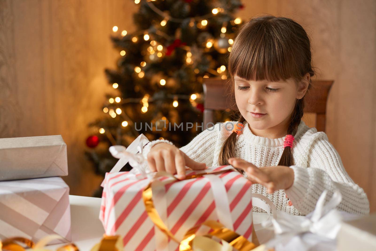 Happy girl opening gift box while sitting at table, female kid with pigtails looking at striped box with interest, little girl posing in room with x,mas tree on background.