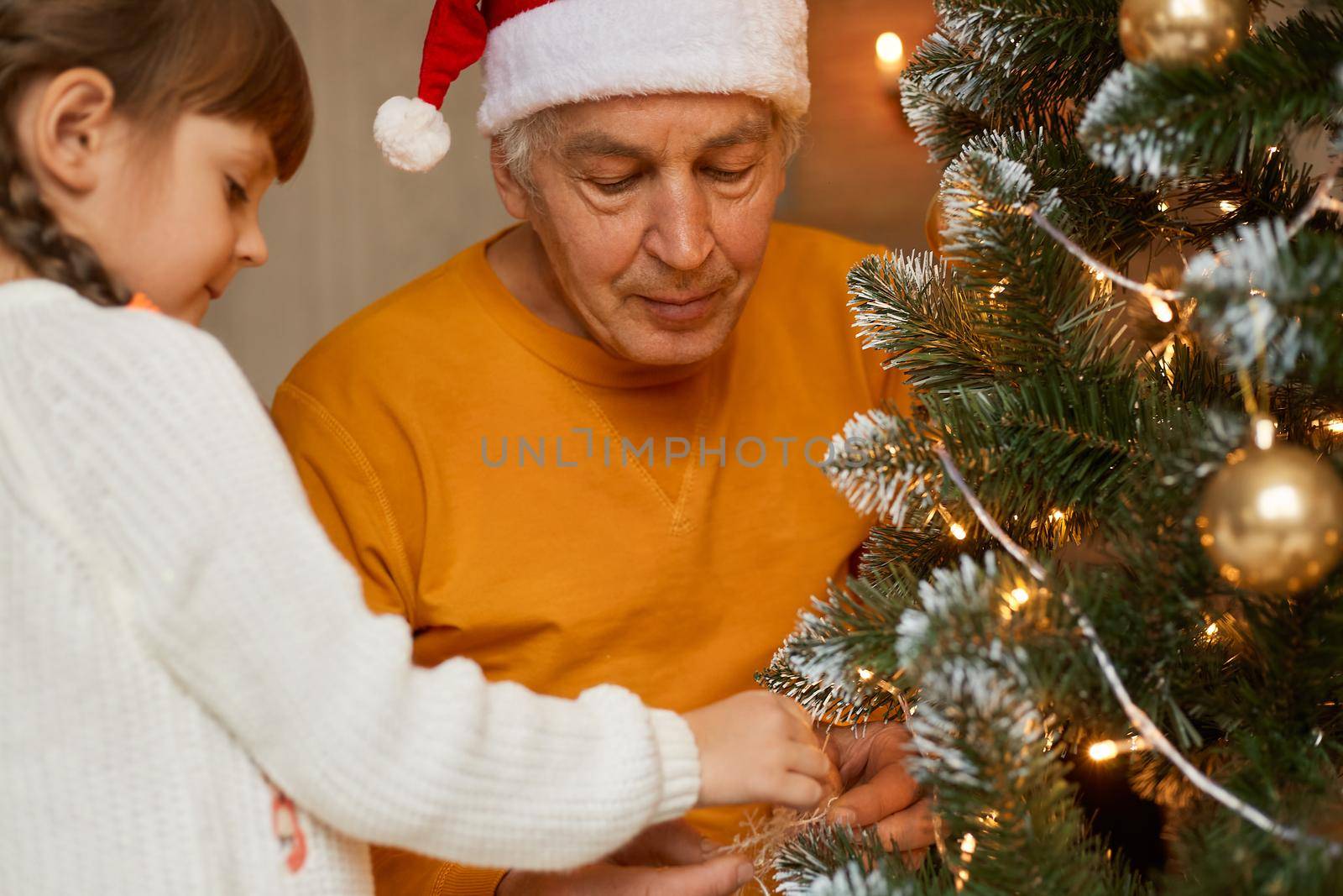 Old man with his grandchild decorating fir tree, posing in festive living room, mature male in orange shirt and santa claus hat looks concentrated at xmas tree, spending time together.