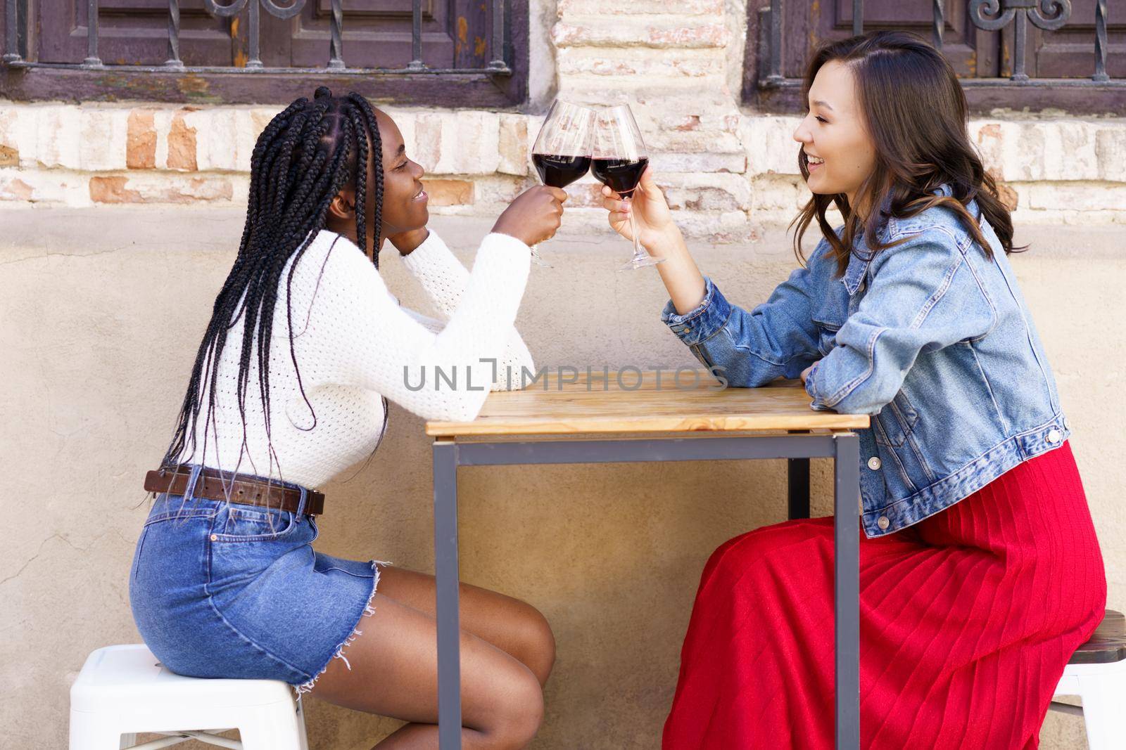 Two women making a toast with red wine sitting at a table outside a bar. by javiindy