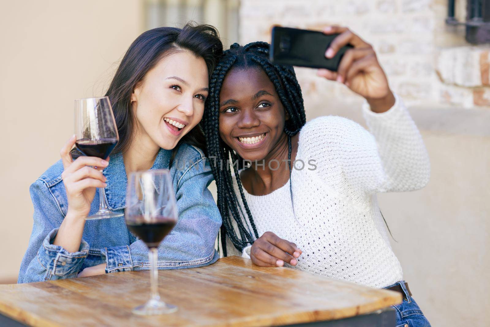 Two friends making a selfie sitting at a table outside a bar while drinking a glass of red wine. by javiindy