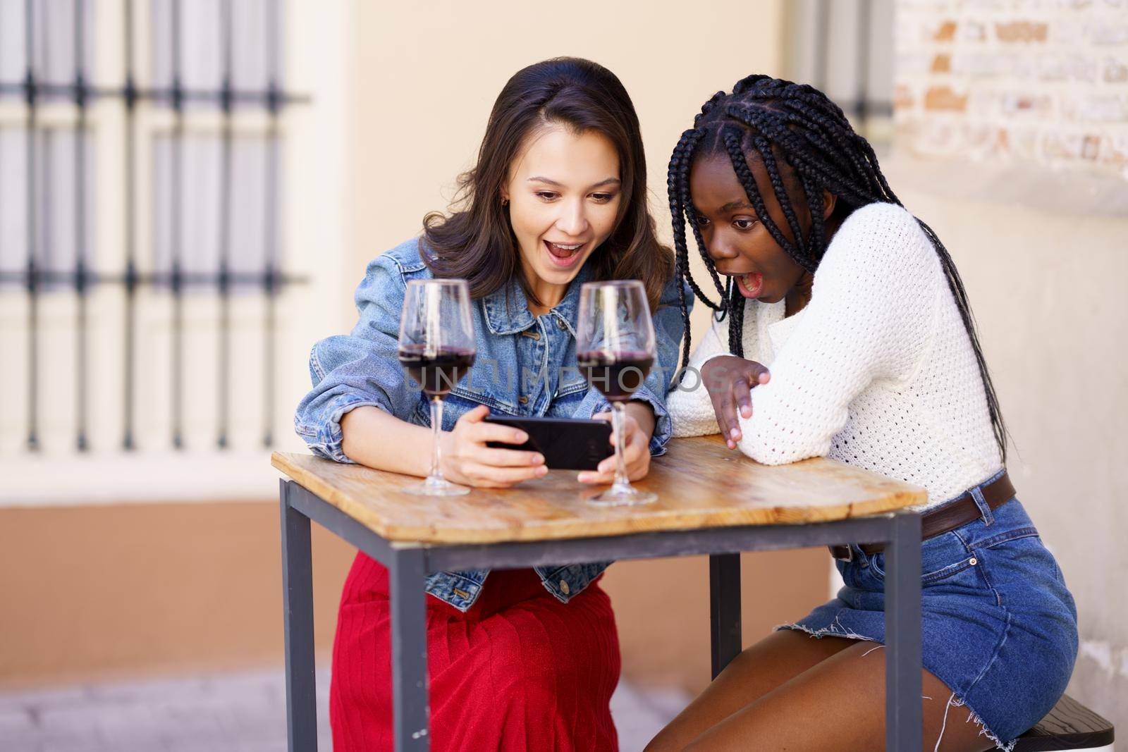 Two women looking at their smartphone together while having a glass of wine. by javiindy