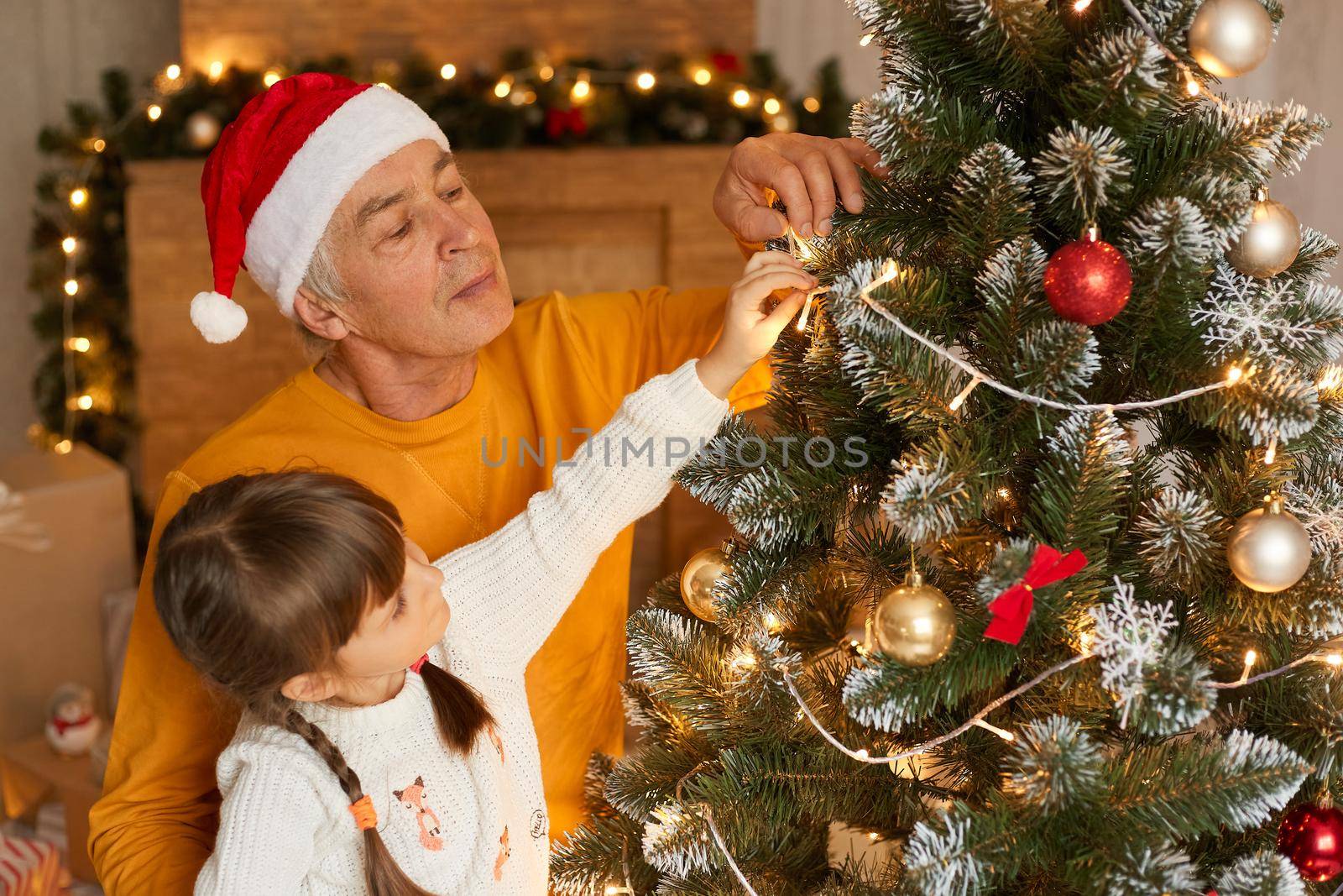 Grandfather and granddaughter decorate fir tree on Christmas eve, looking concentrated at xmas tree, wearing casual attires, posing in festive living room, feels happy to spend time together.