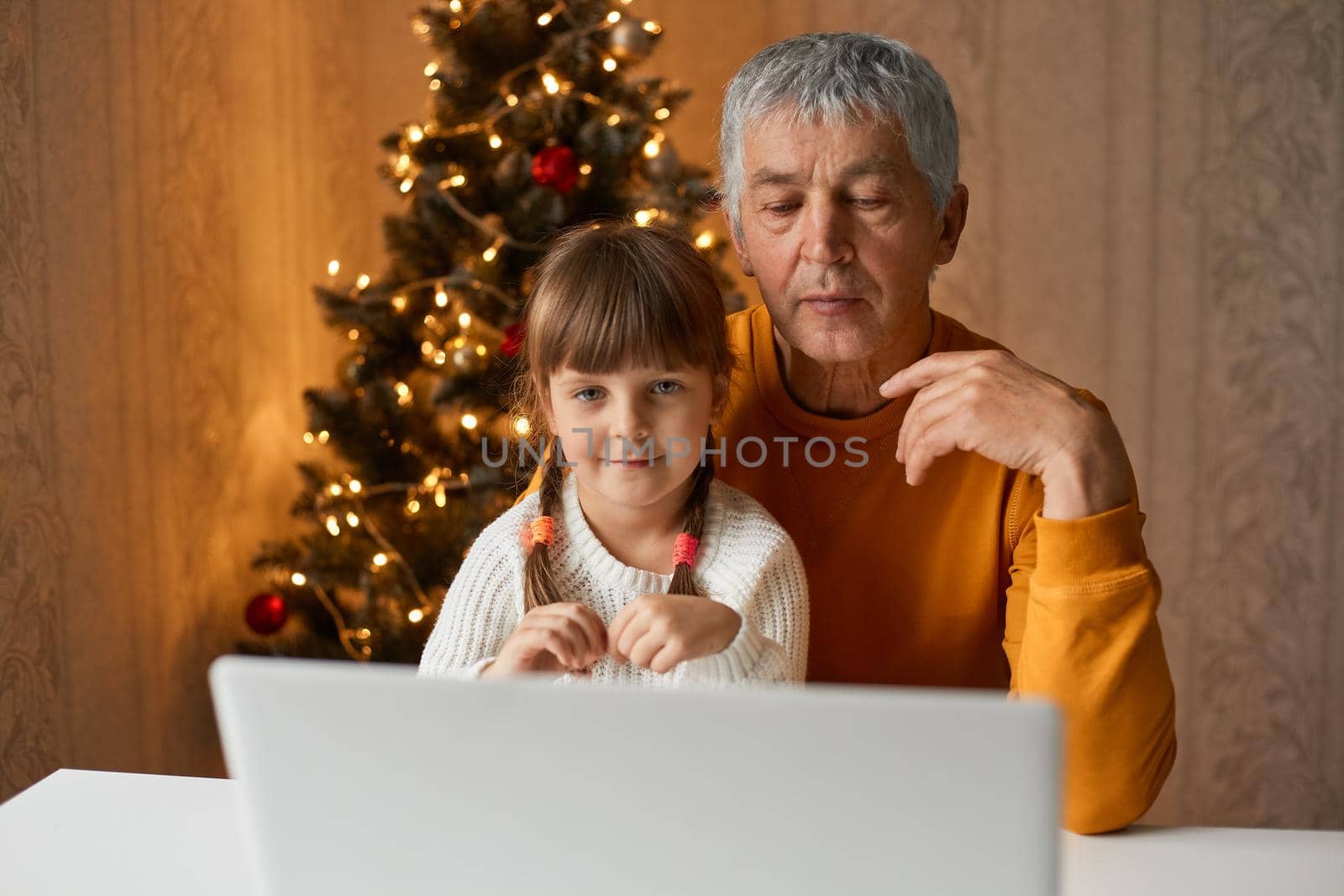 Elderly man with little female child talking to somebody via video call, keeping distance, during COVID-19. Family posing at home having celebration New year.