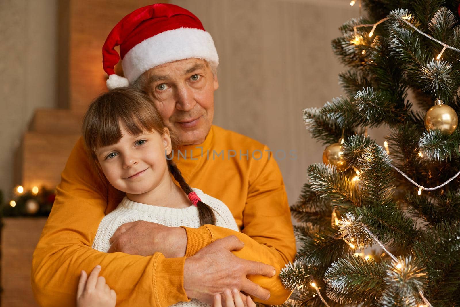 Happy family on Christmas eve, senior man hugging little charming girl with pigtails dresses white shirt, grandpa with grandchild posing in rom with new year decoration.