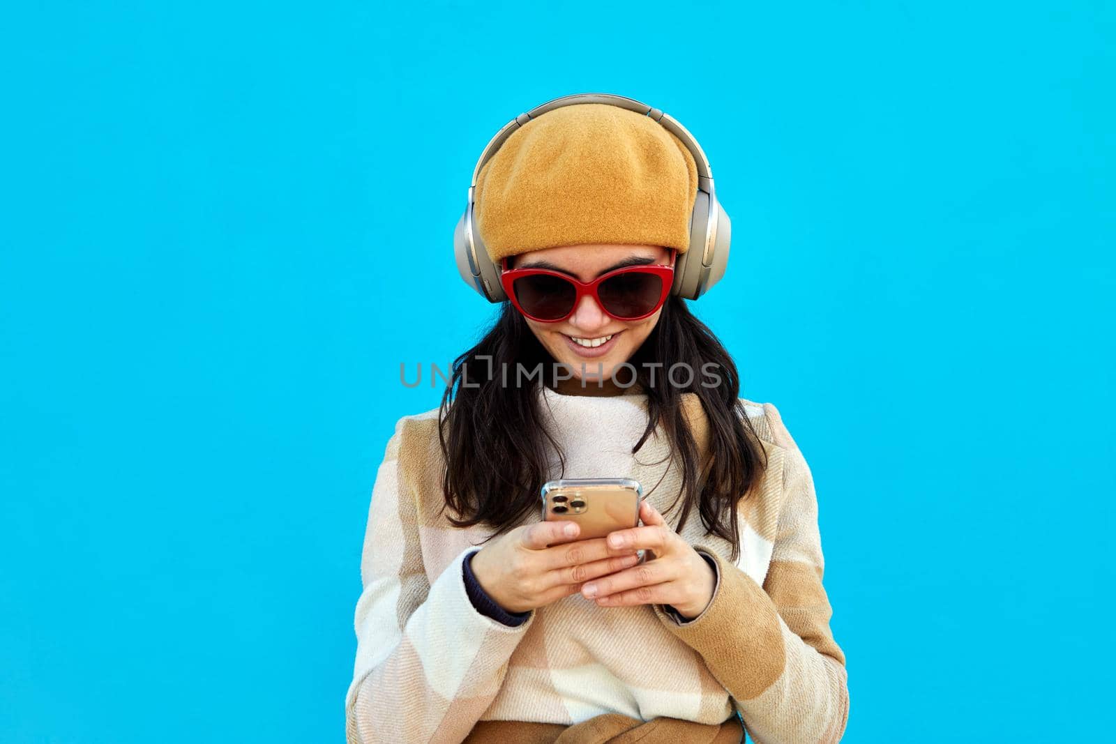 Positive female with black hair in sunglasses and hat listening to music in headphones while surfing cellphone on blue background