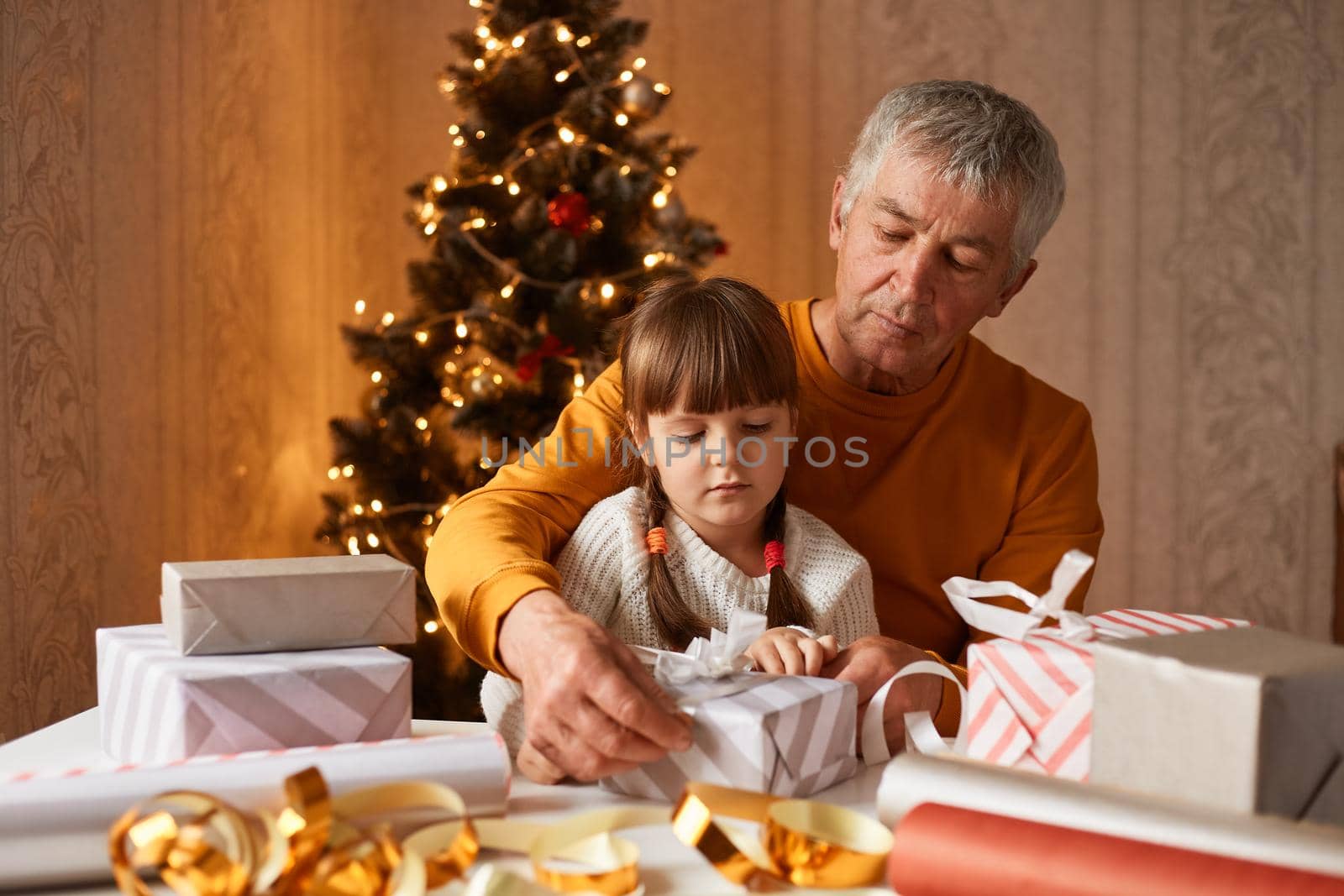 Indoor shot of senior man wearing orange sweater with his granddaughter on knees packing present boxes for Christmas holidays while sitting at table together.