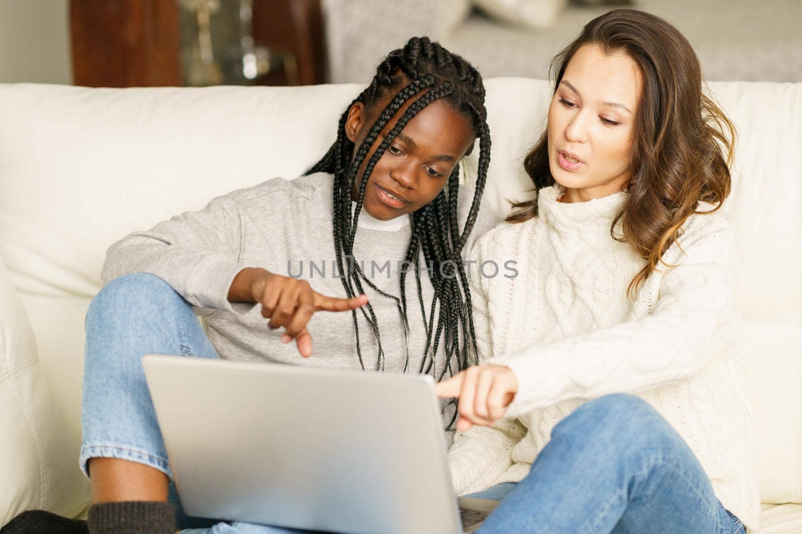 Two female student friends sitting on the couch at home using a laptop. Multiethnic women.