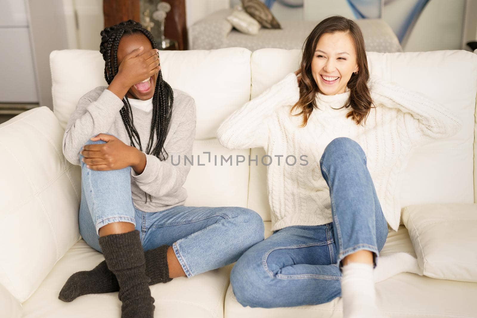 Two female student friends laughing together on the couch at home. Multiethnic women.