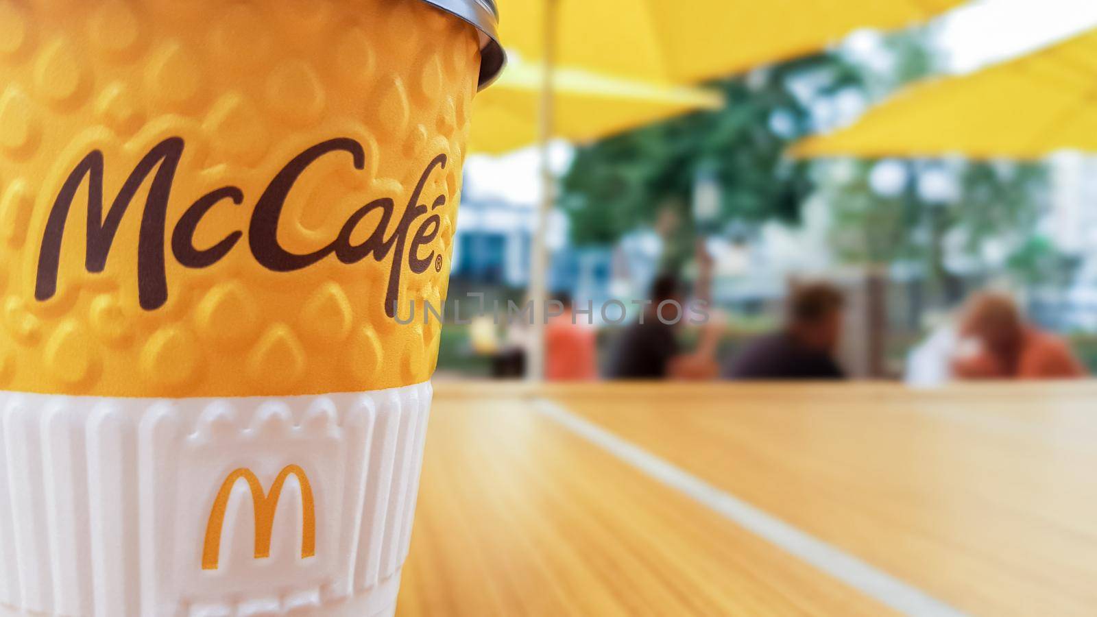 McCafe menu at McDonalds restaurant. Yellow cup of coffee on a table outdoors with yellow umbrellas and visitors in the background. Breakfast, coffee time. Ukraine, Kiev - September 6, 2019