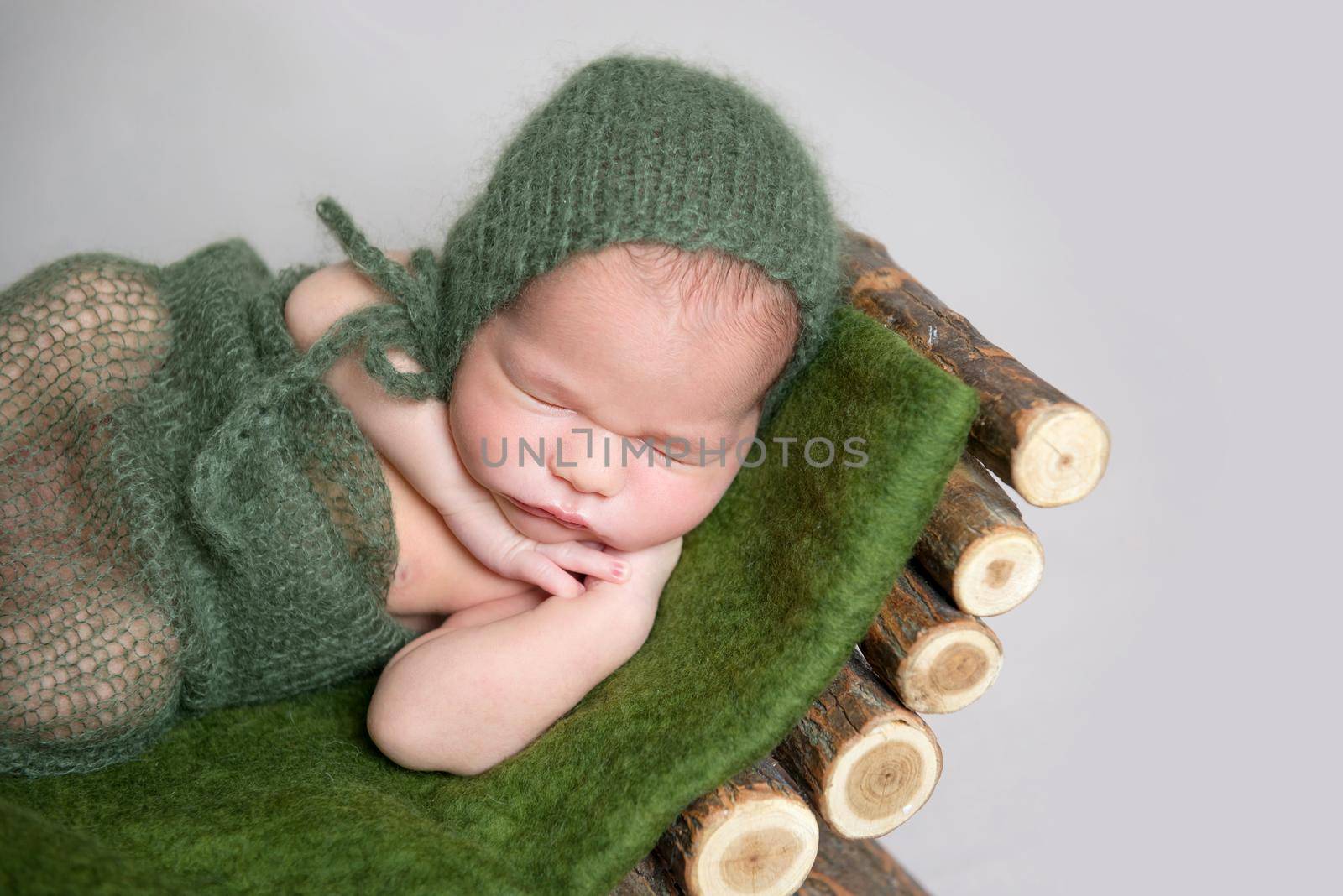 Cute sleeping baby wrapped in green diaper and knitted hat on small wooden crib.