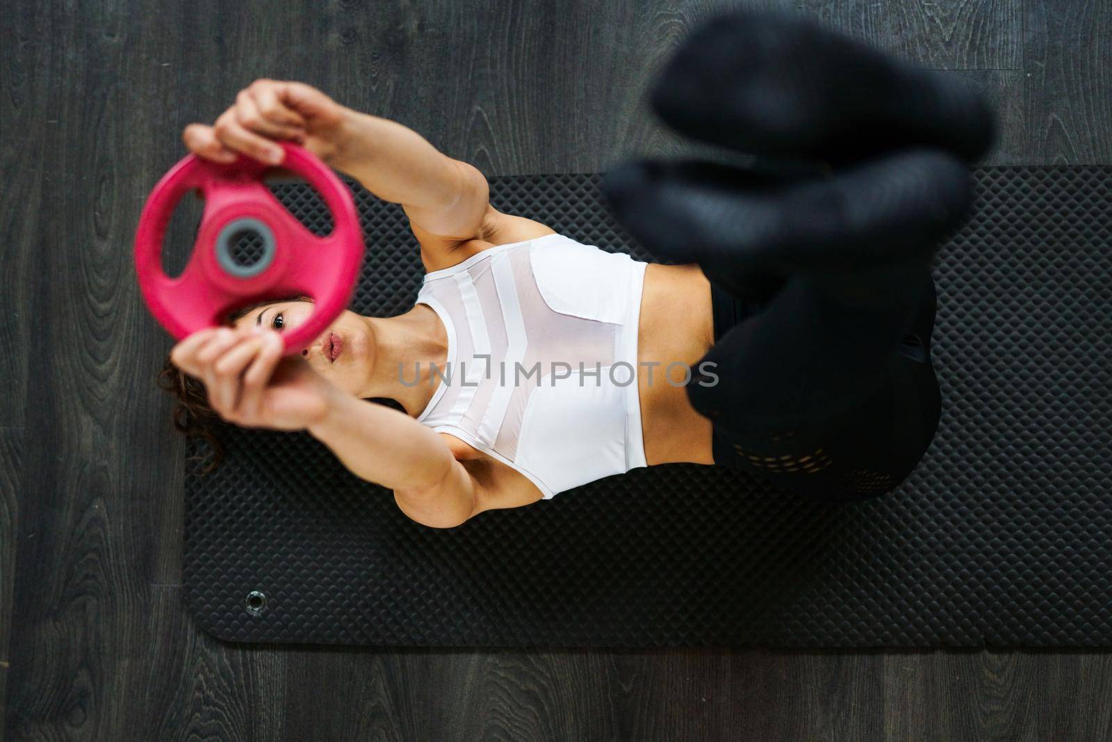 Sporty young female, wearing sportswear, on yoga mat doing situps with plate weights in gym. Fitness woman doing abs crunches.