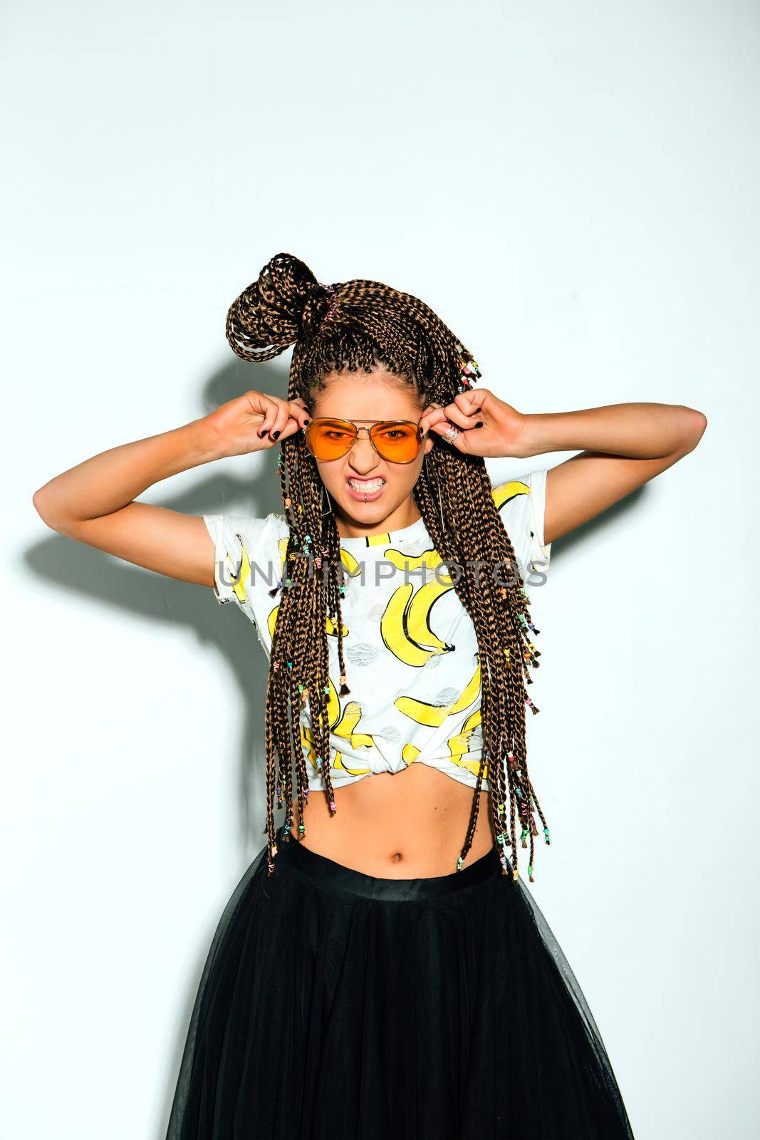 Portrait of a young stylish woman with braided hair dressed in white t-shirt, black skirt and yellow sunglasses on the white background.