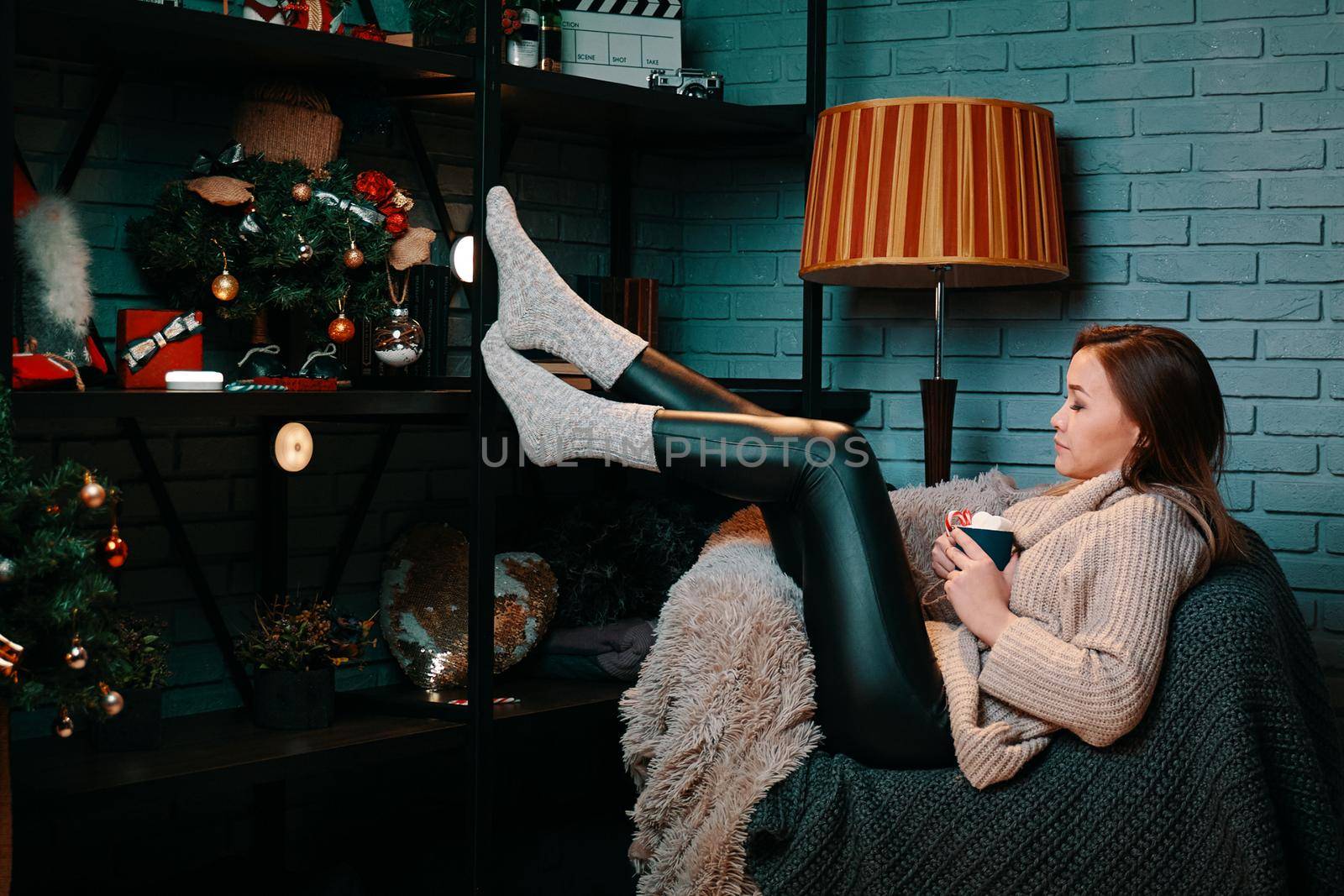 Cute woman with cup of coffee sitting on chair. Hot chocolate with marshmallows. Christmas tree and decorations on shelf. New year's mood. Knitted blankets on chair. Gray brick wall on background.