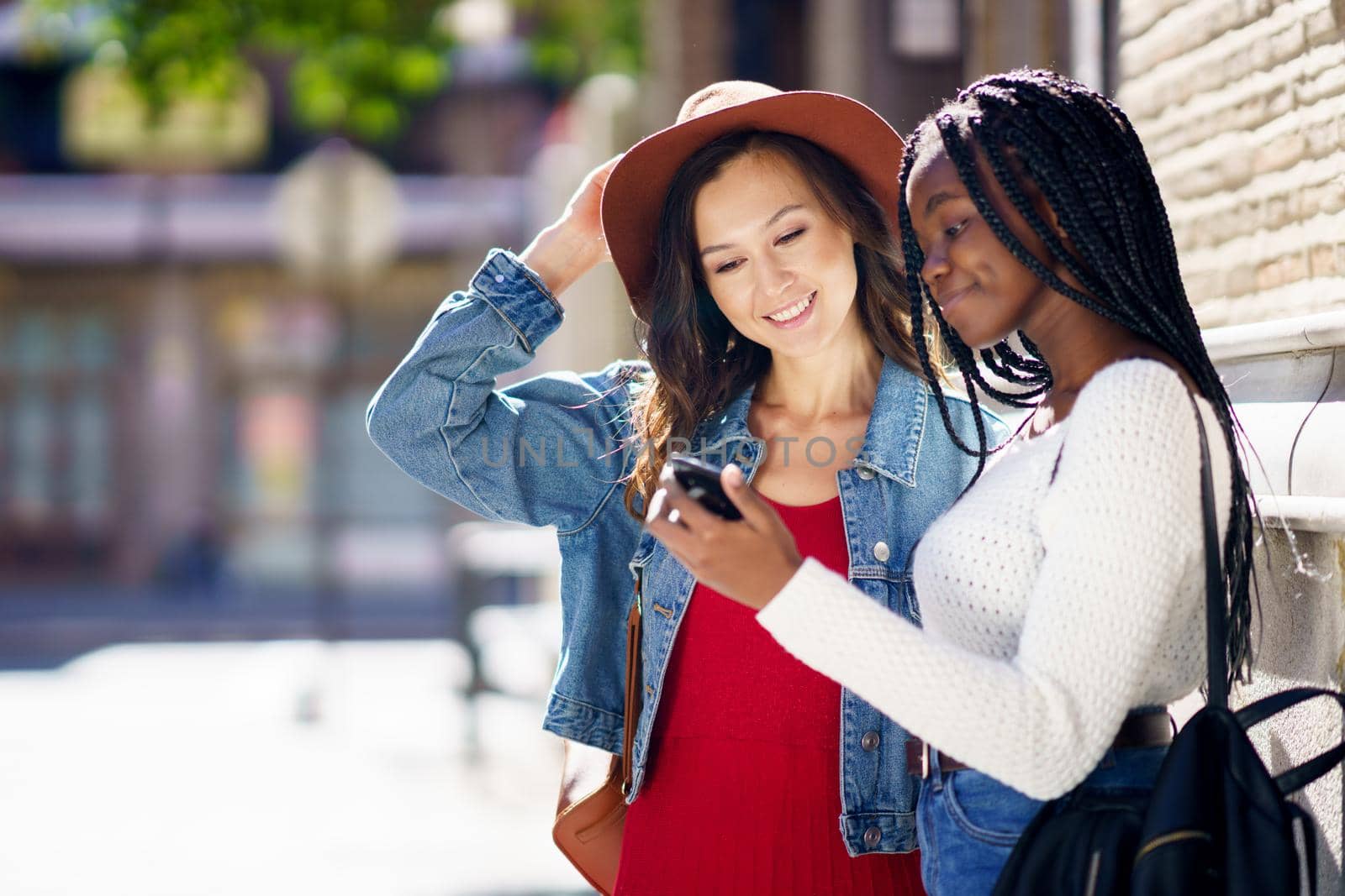 Two friends looking at their smartphone together on the street. Multiethnic women.