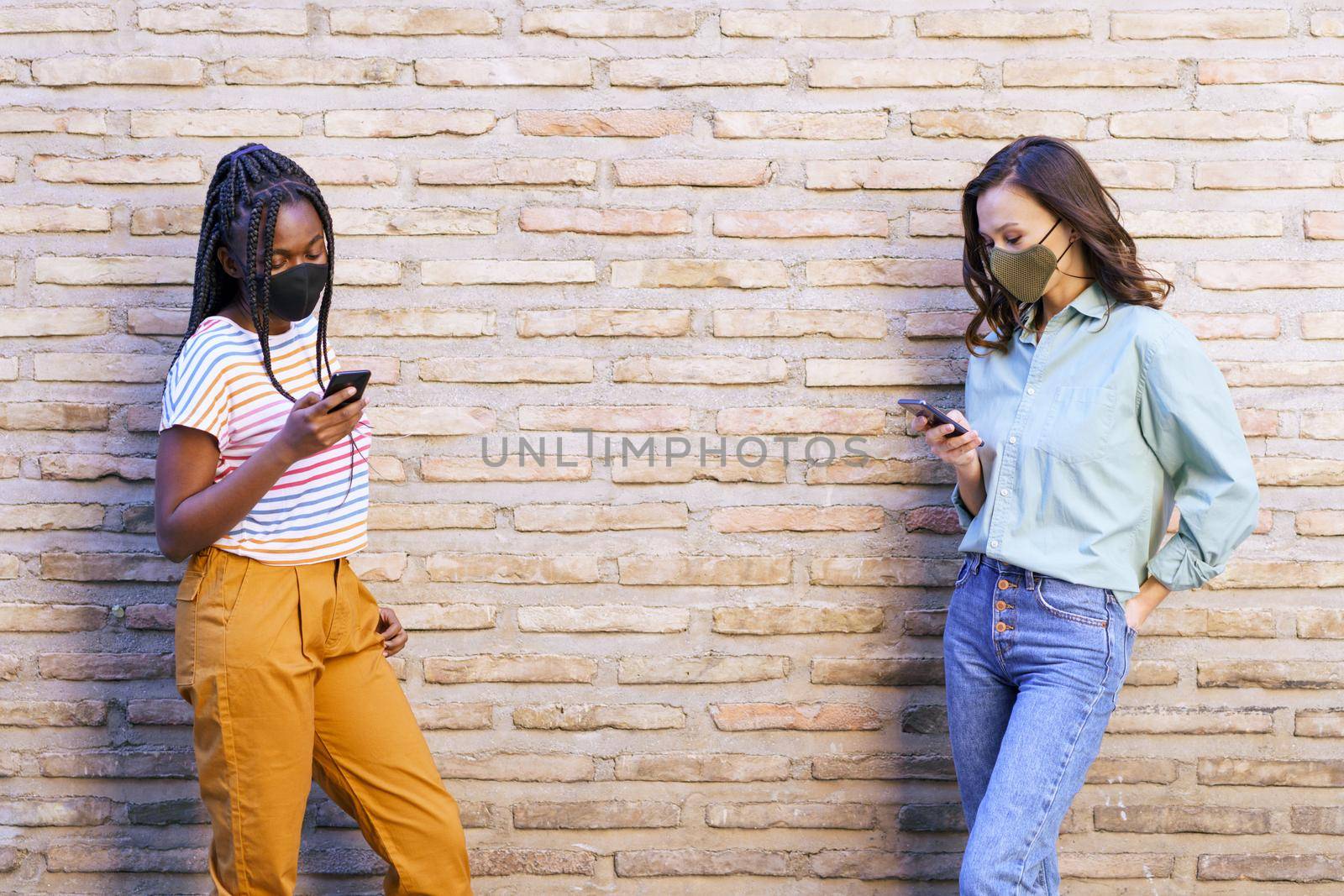Multiethnic young women wearing masks using smartphone separated to respect social distance outdoors.