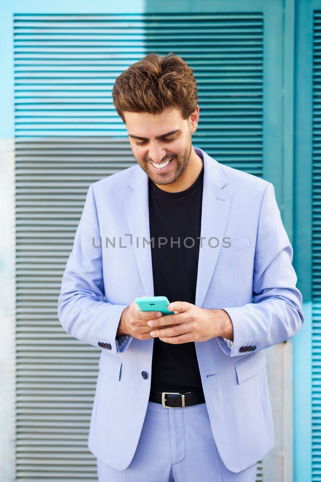 Smiling man wearing blue suit texting with a smartphone in urban background.