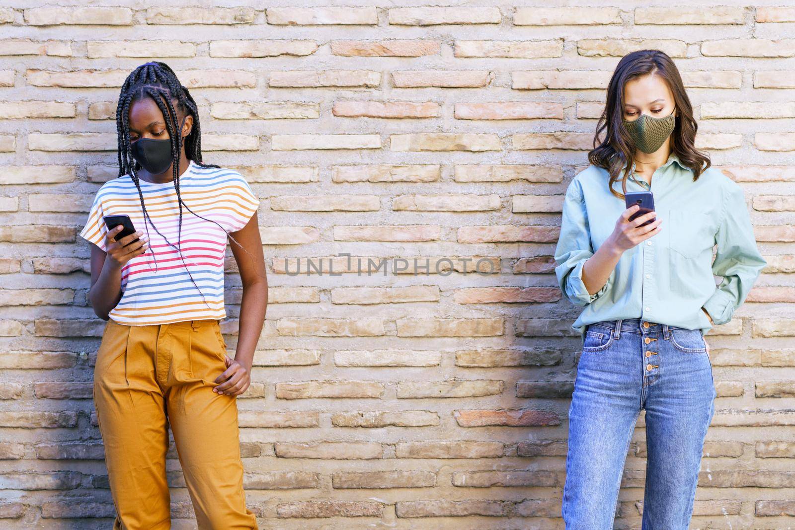 Multiethnic young women wearing masks using smartphone separated to respect social distance. by javiindy