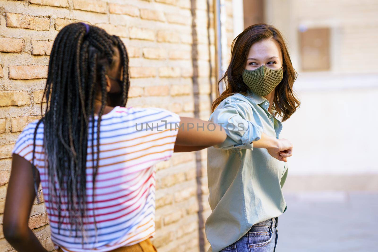 Multiethnic young women wearing masks greeting at each other with their elbows outdoors