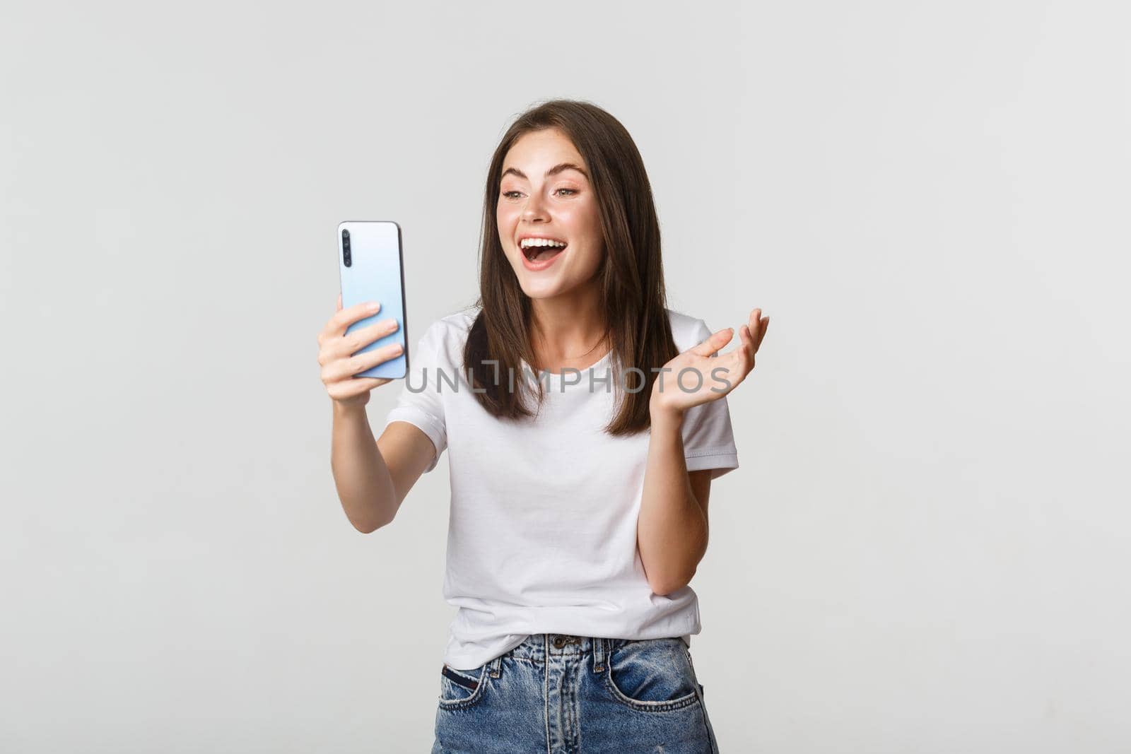 Friendly smiling woman video calling and talking on smartphone happy, standing white background.