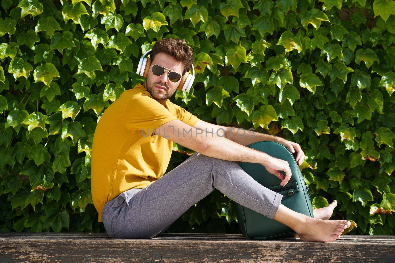 Young student sitting outside wearing headphones and backpack. by javiindy