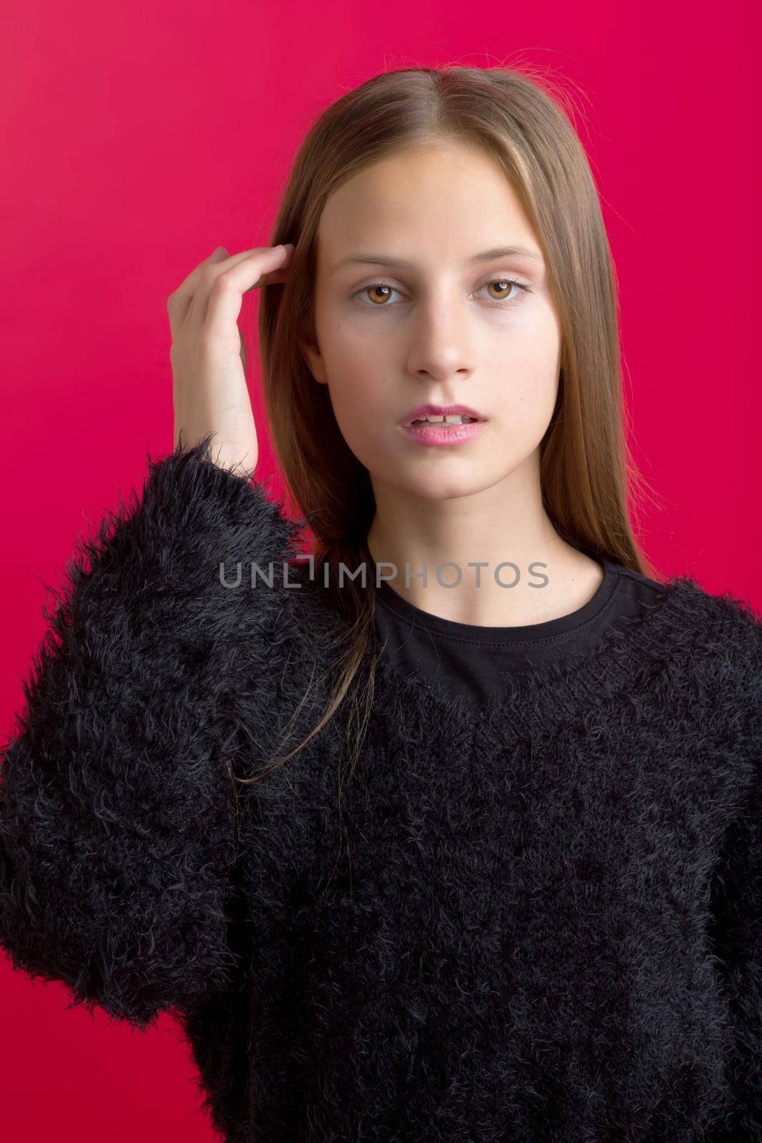 Beautiful teenage girl touching her long hair. Portrait of charming smiling teenager wearing black stylish oversized sweater with leather belt standing on red background