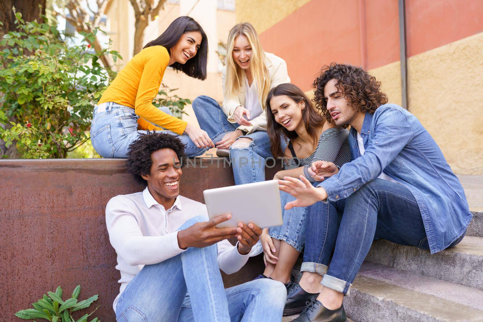 Young black man showing something on his digital tablet to his group of friends sitting on some steps in the street.