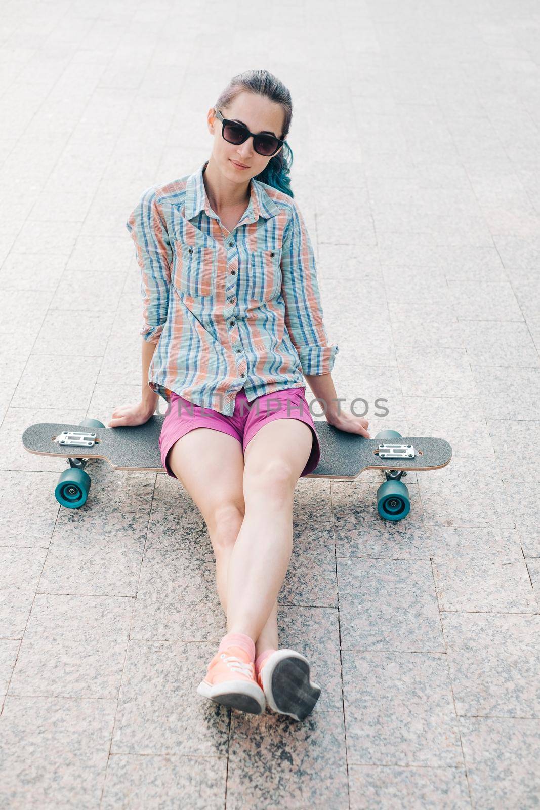 Smiling girl in sunglasses sitting on longboard, looking at camera.