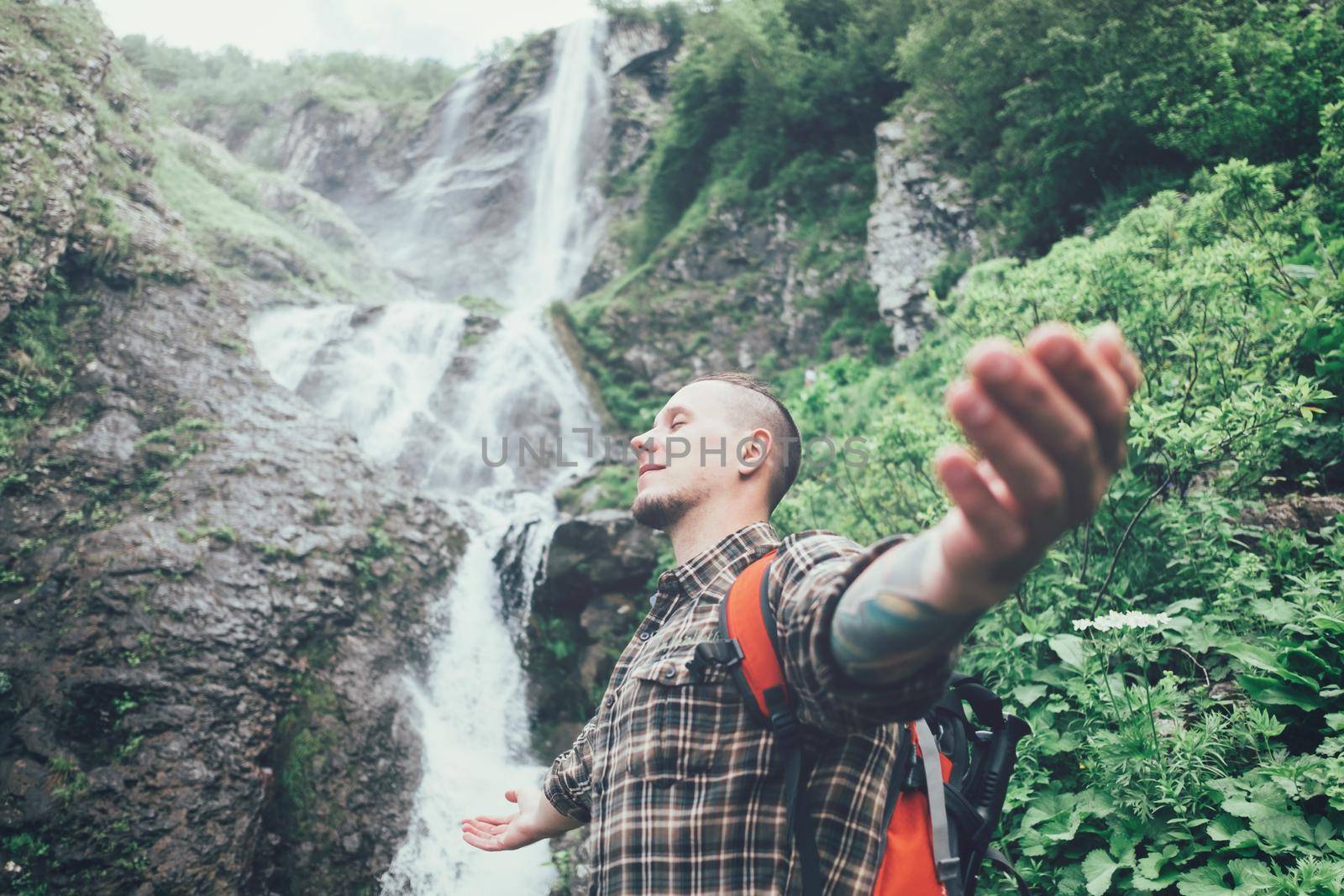 Happy backpacker young man standing with raised arms on background of waterfall and enjoying of summer nature, side view.