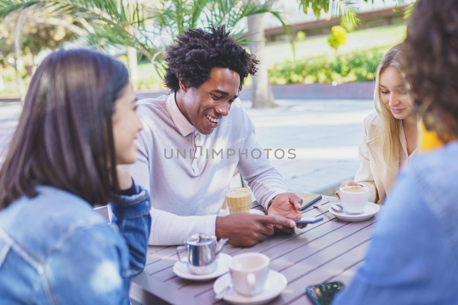 Multi-ethnic group of friends having a drink together in an outdoor bar. One of the men shows something on his smartphone to his friends.