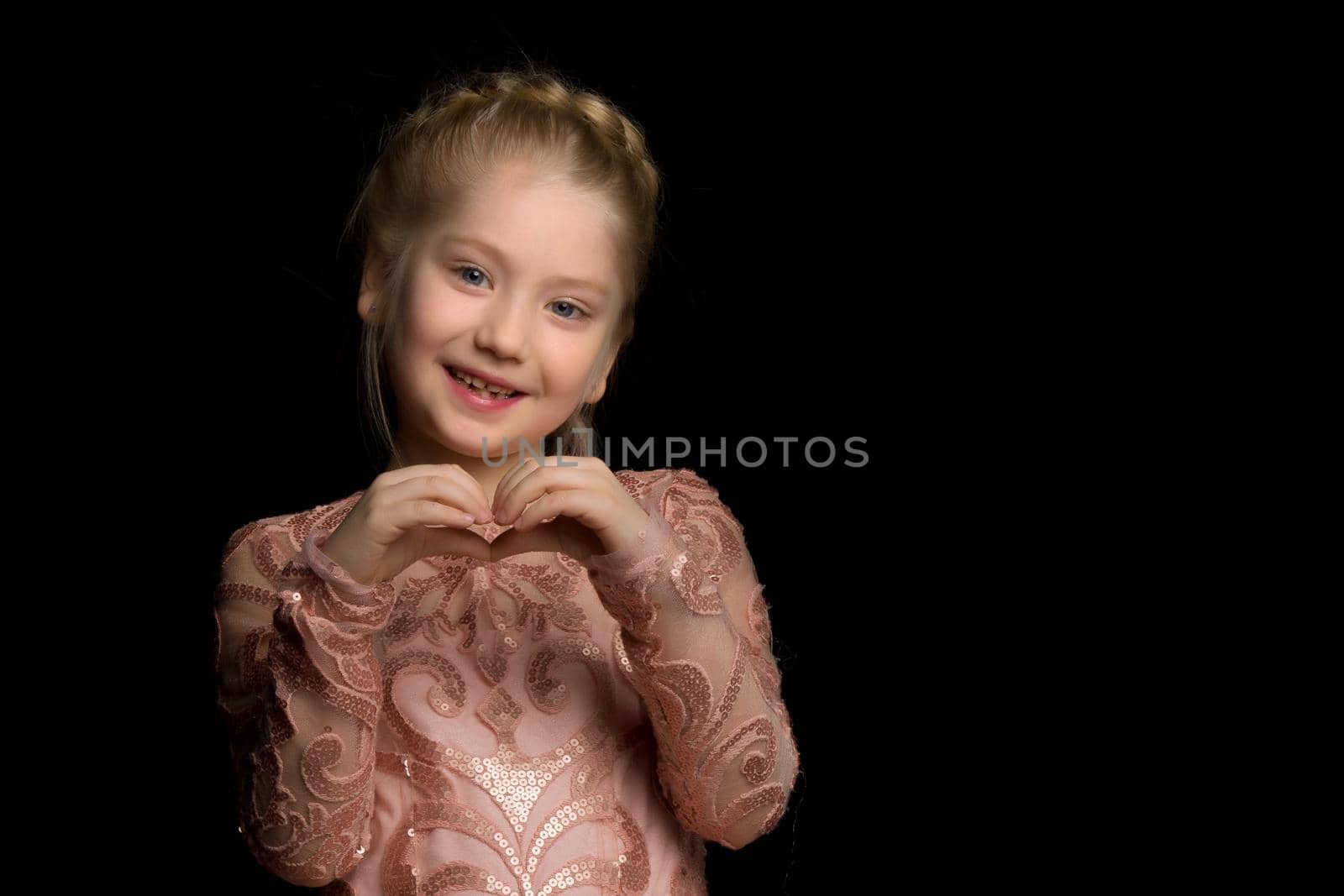 Cute little girl makes a heart with her hands and smiles on a black background.