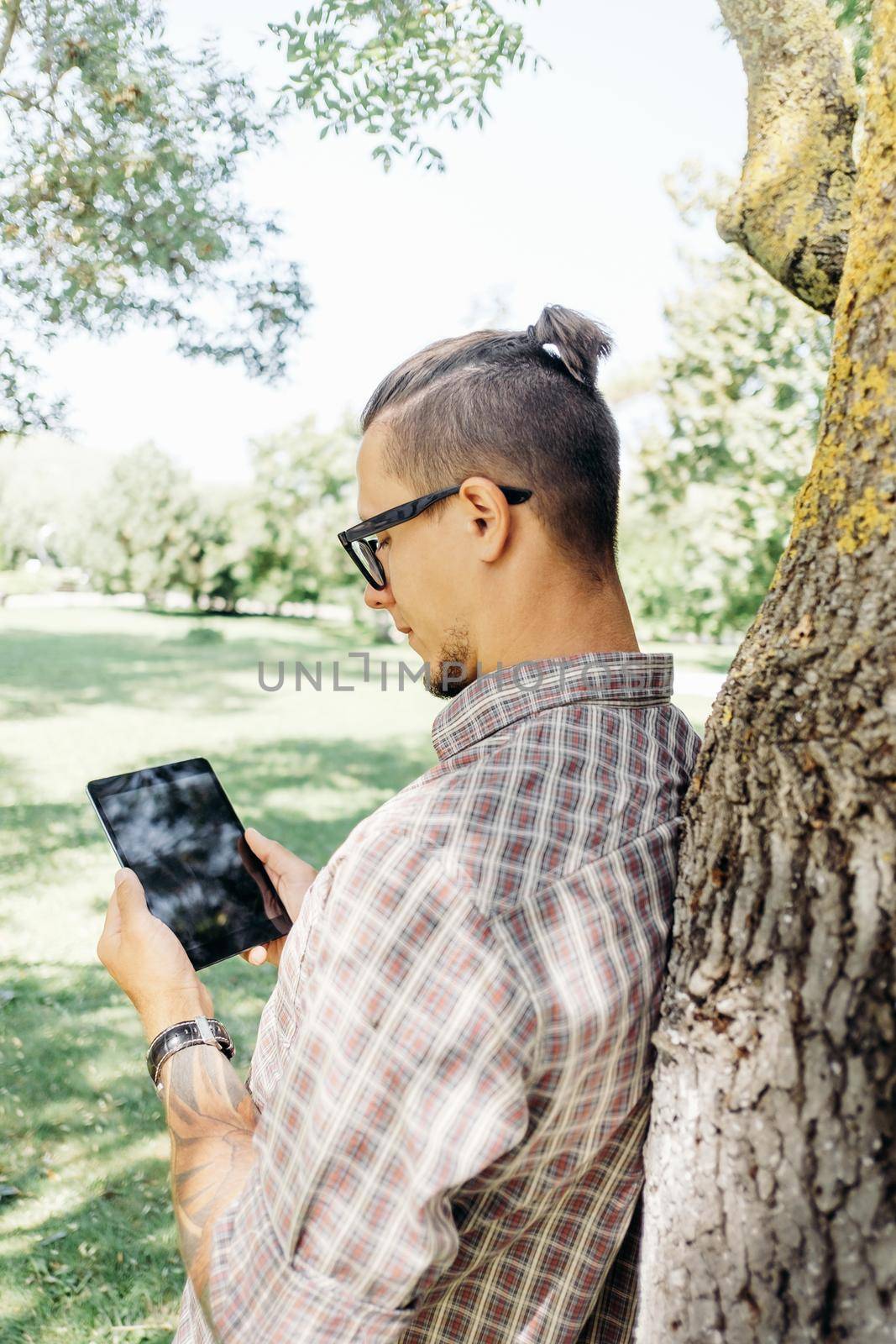 Stylish young man standing near a tree with digital tablet in summer park.