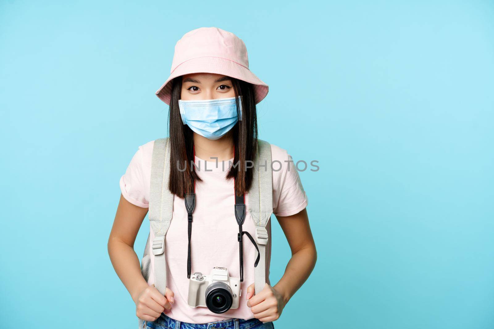 Happy asian girl tourist, standing in medical face mask, holding camera, taking pictures during vacation, sightseeing, explore abroad, standing over blue background.
