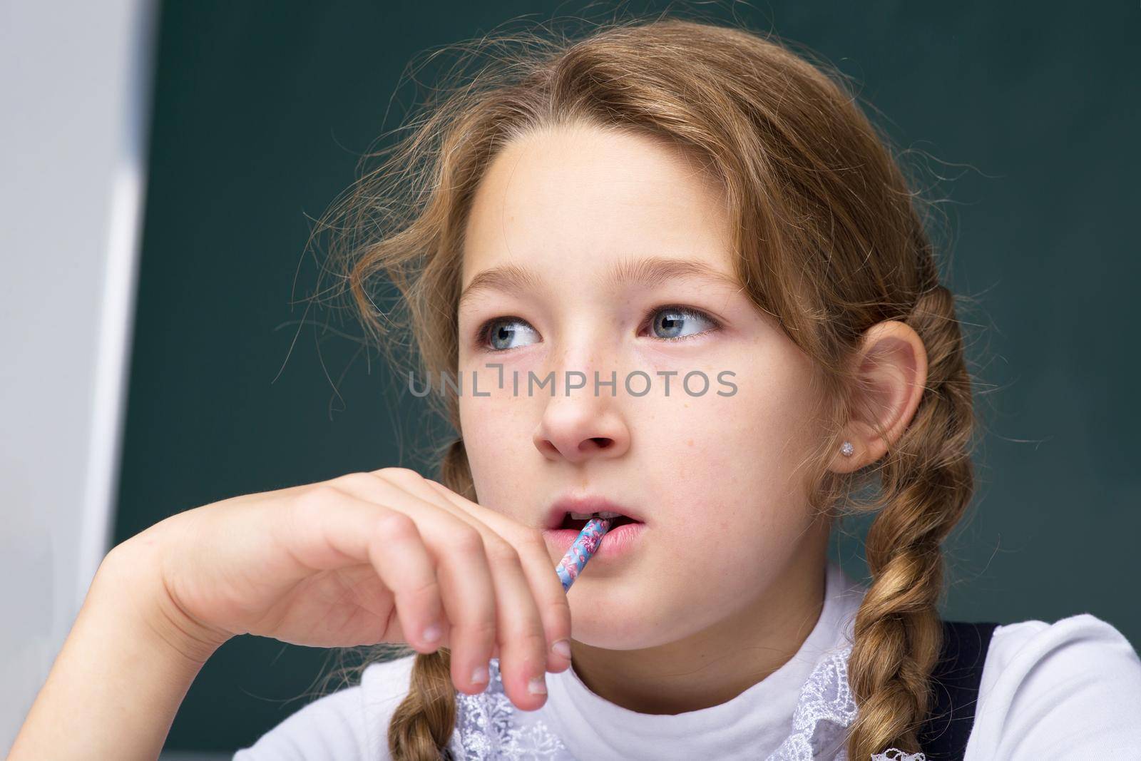 Portrait of pensive schoolgirl. Thoughtful girl holding pen near her mouth looking away. Primary school student posing on background of blackboard. Back to school, education concept