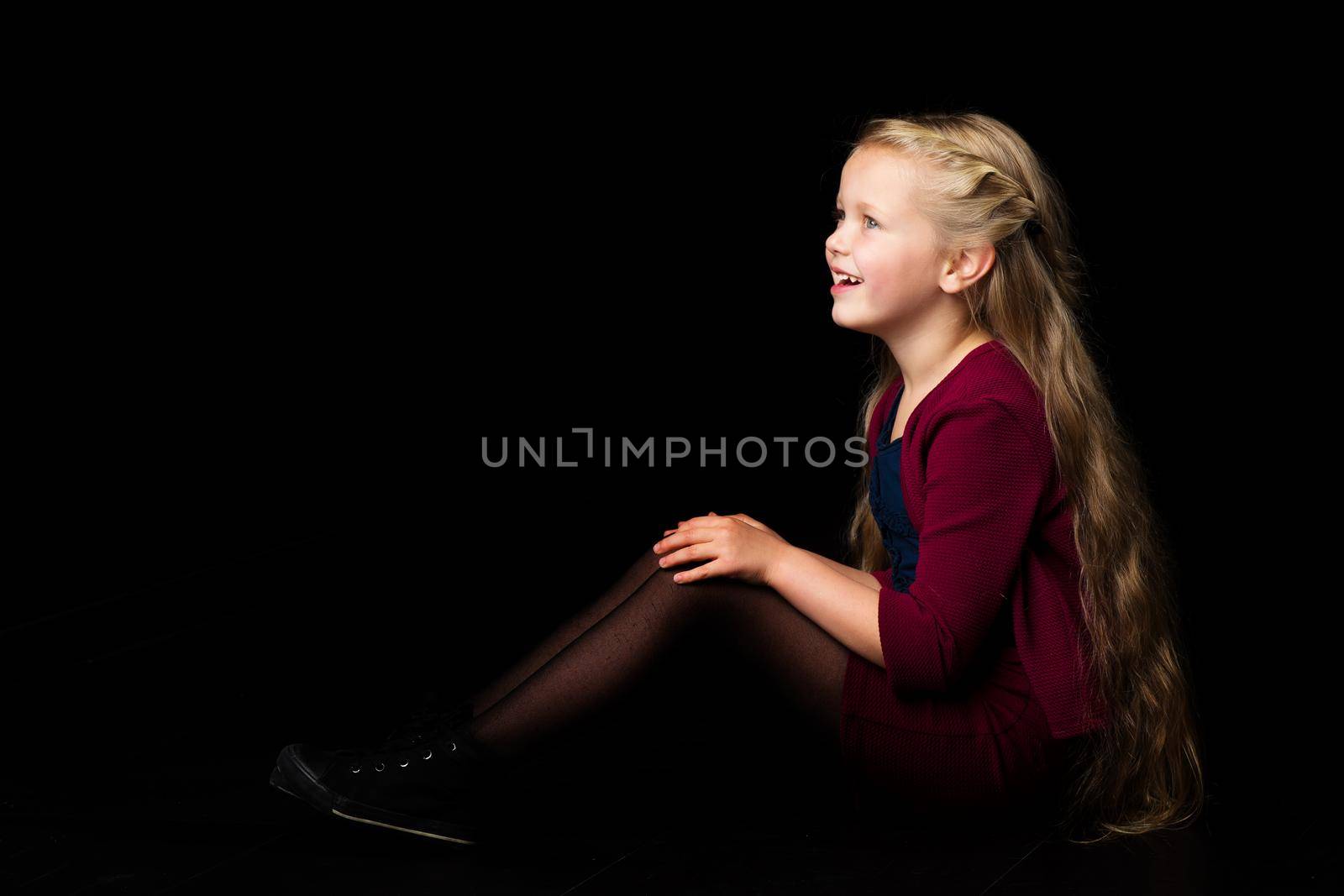 Side view of happy smiling blonde girl. Happy long haired girl in stylish clothes sitting on floor with her knees up. Lovely little child posing in studio against black background