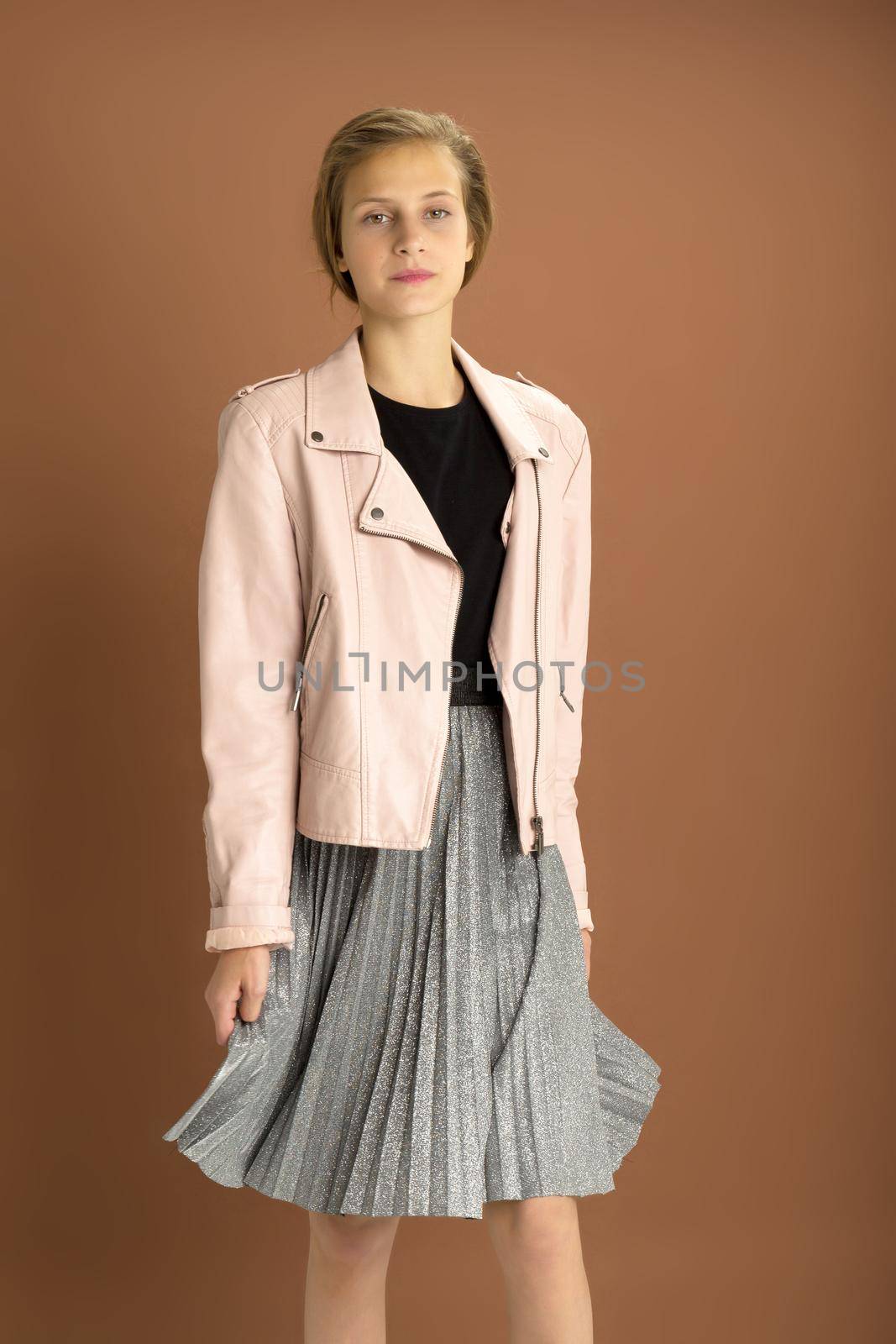 Happy girl whirling against brown background. Joyful attractive brown haired teen girl dressed pink leather jacket and gray fluttering pleated skirt having fun in studio
