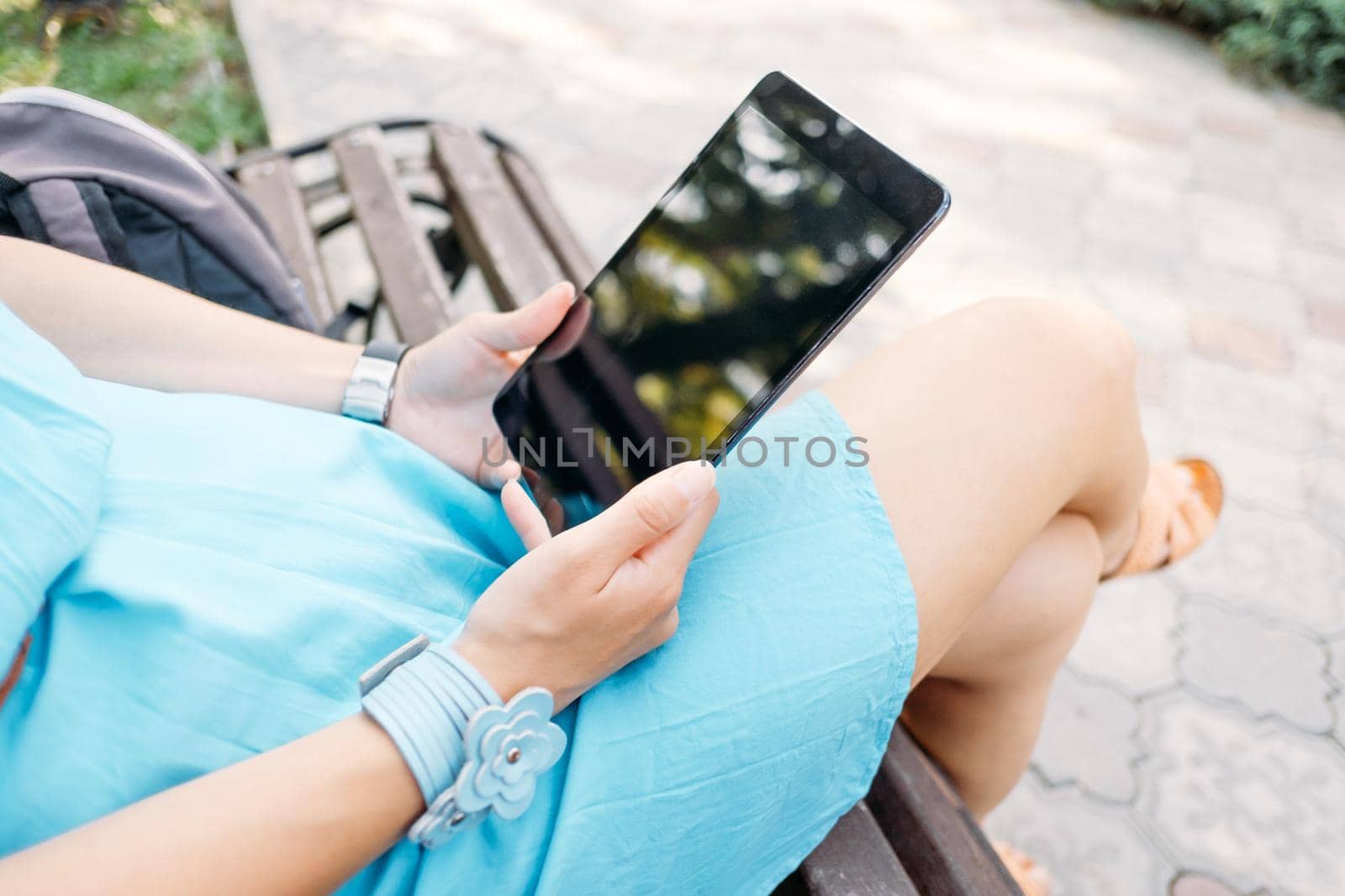 Unrecognizable young woman sitting with digital tablet on wooden bench in the park.