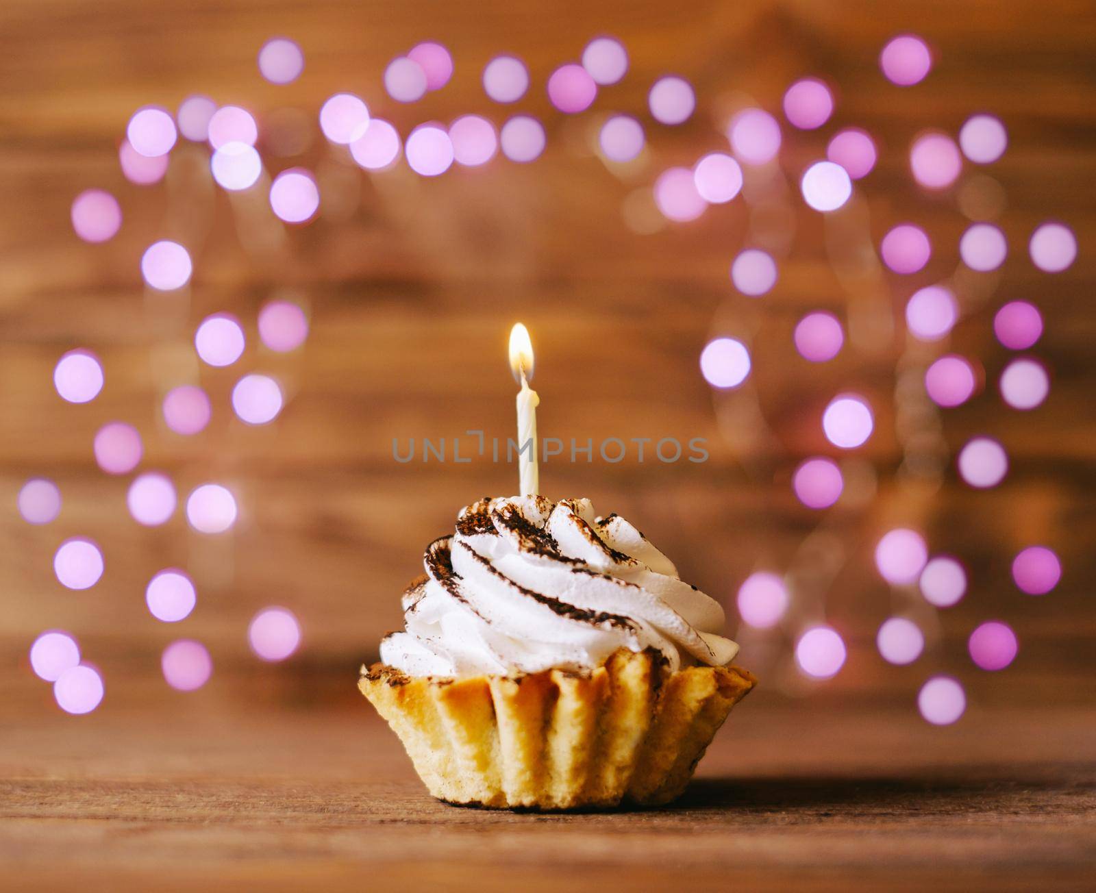 Birthday cream cupcake with candle on a wooden table against blurred lights.