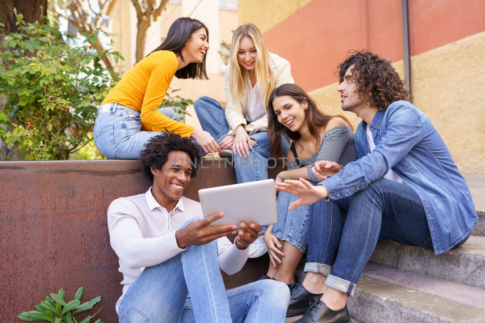 Multi-ethnic group of young people looking at a digital tablet outdoors in urban background. Group of men and woman sitting together on steps.