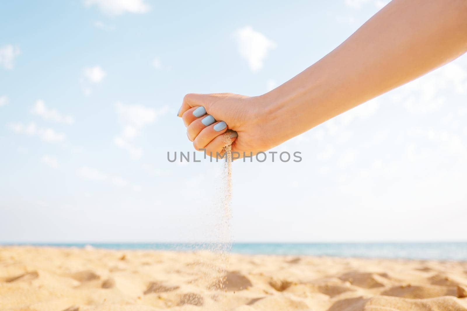 Female hand pours sand on beach near the sea on background of blue sky.