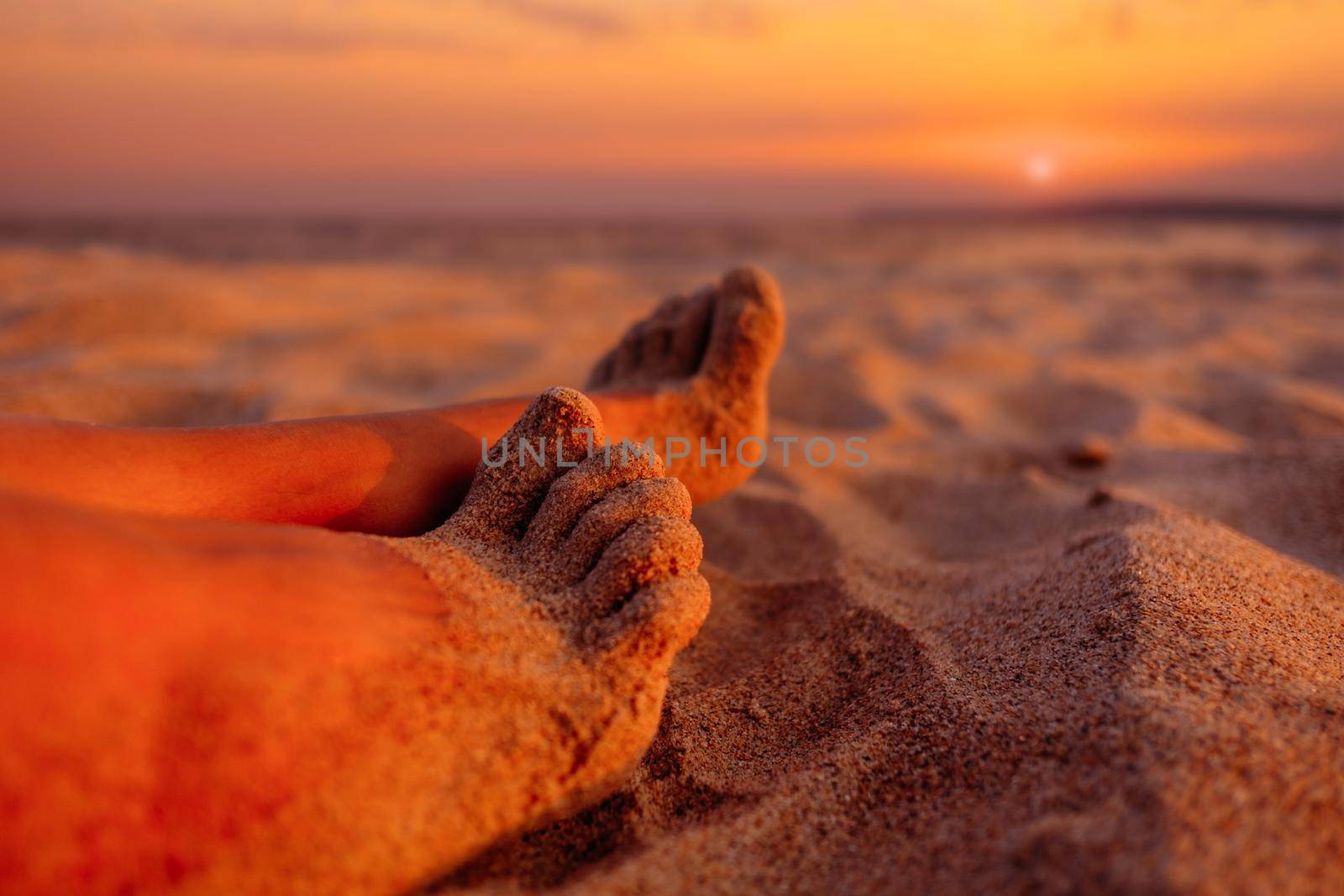 Woman resting on beach near the sea, close-up view of female barefoot legs sand at sunset.