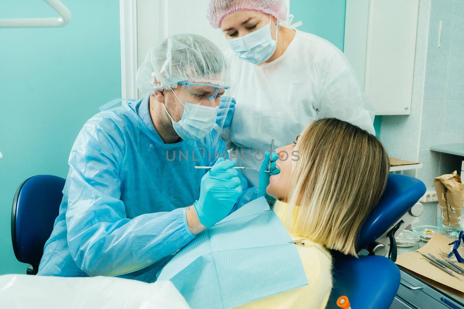 a dentist in a protective mask sits next to him and treats a patient in the dental office with an assistant by Lobachad