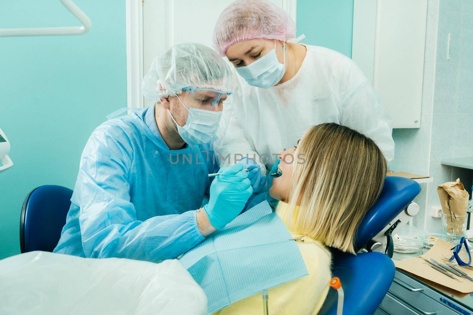 a dentist in a protective mask sits next to him and treats a patient in the dental office with an assistant.