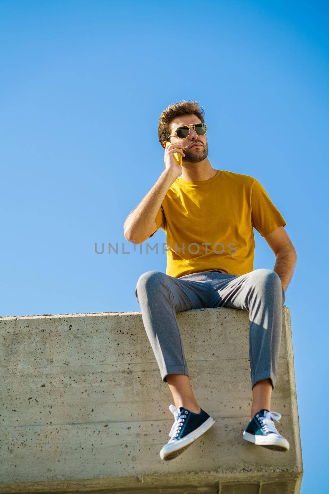 Young man using his smartphone sitting on a ledge outside by javiindy