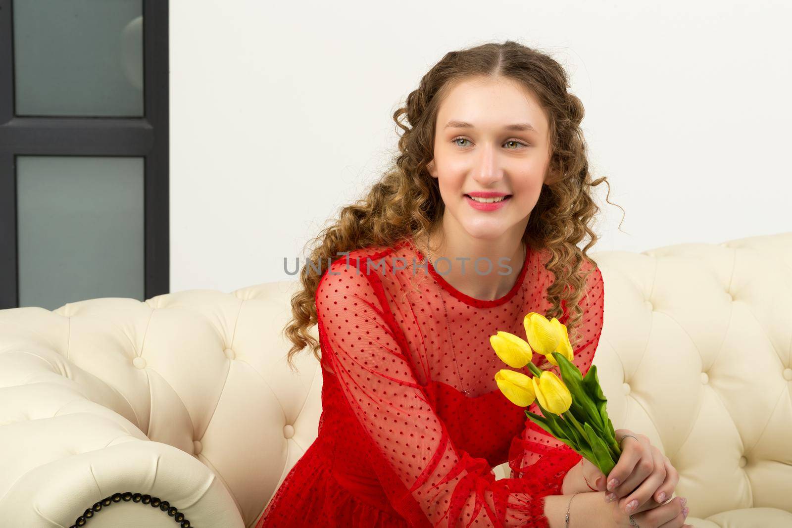 Portrait of pretty girl with fresh yellow tulips. Cheerful happy blonde young woman in red dress posing with bouquet of flowers on light background in studio