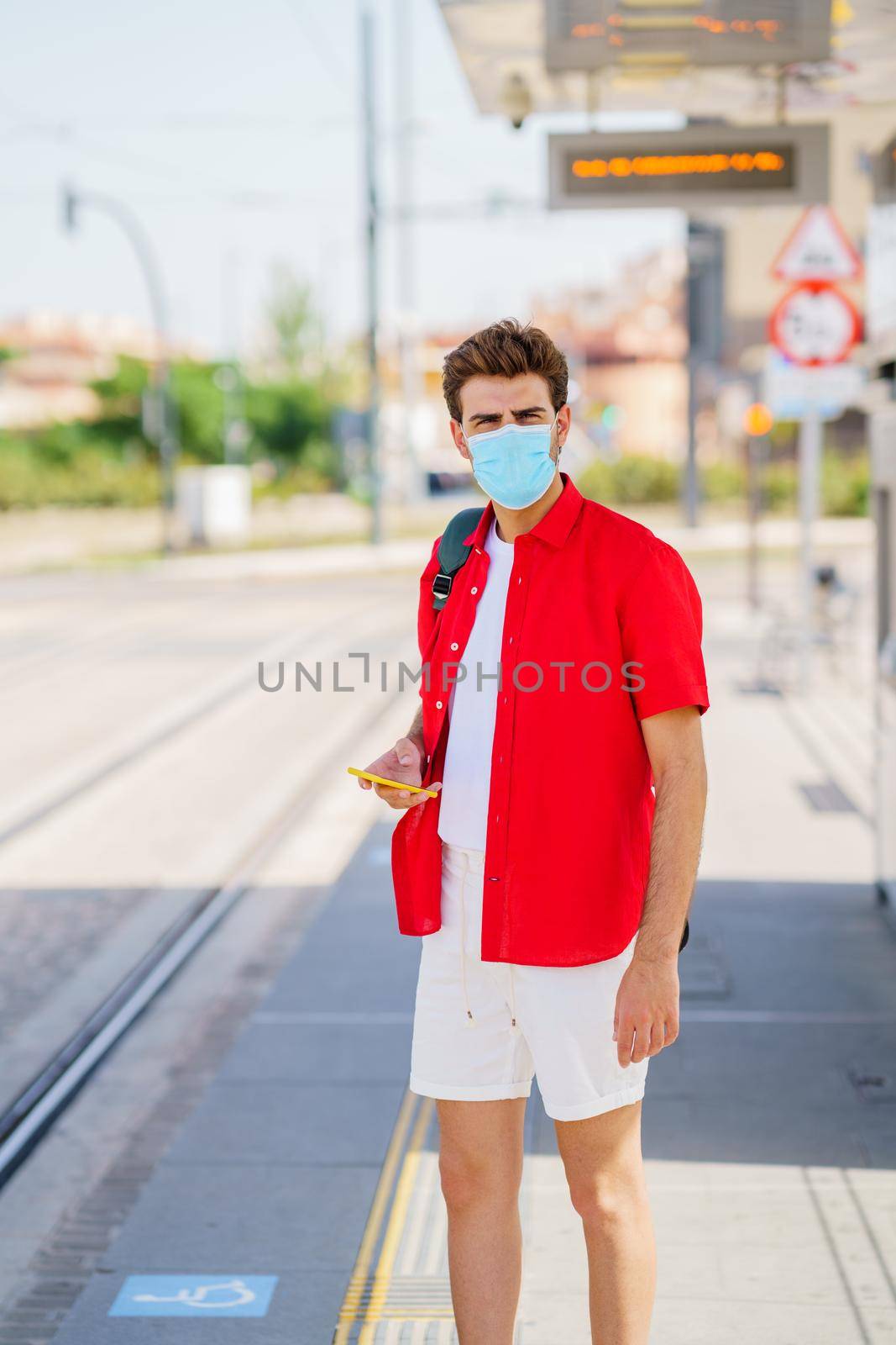 Young man wearing a surgical mask while waiting for a train at an outside station by javiindy