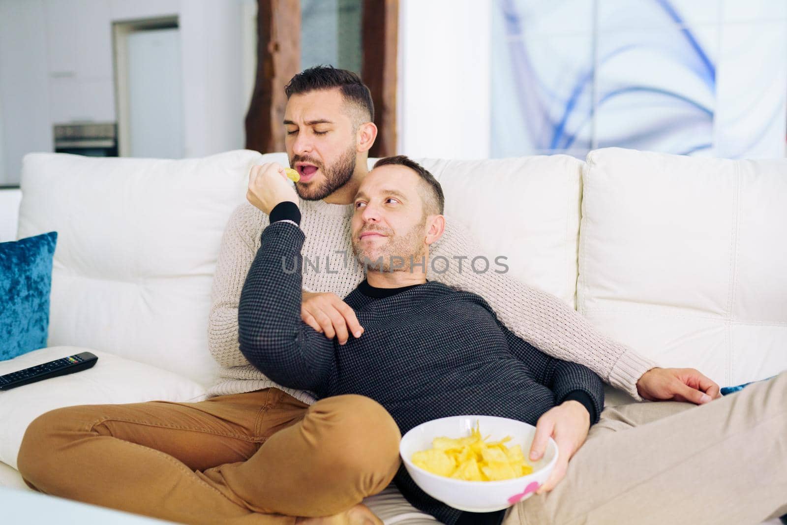 Gay couple sitting on the couch at home watching something on TV and having a snack. Homosexual relationship concept.
