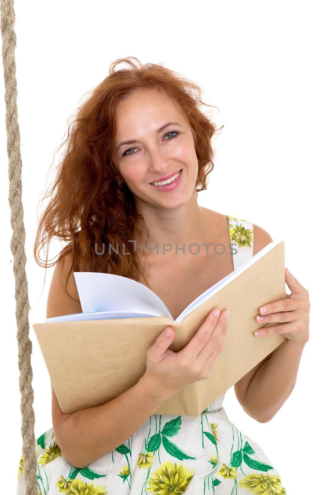 Happy young woman reading book on swing. Smiling woman wearing summer dress sitting on rope swing against white background. Portrait of beautiful girl posing in studio