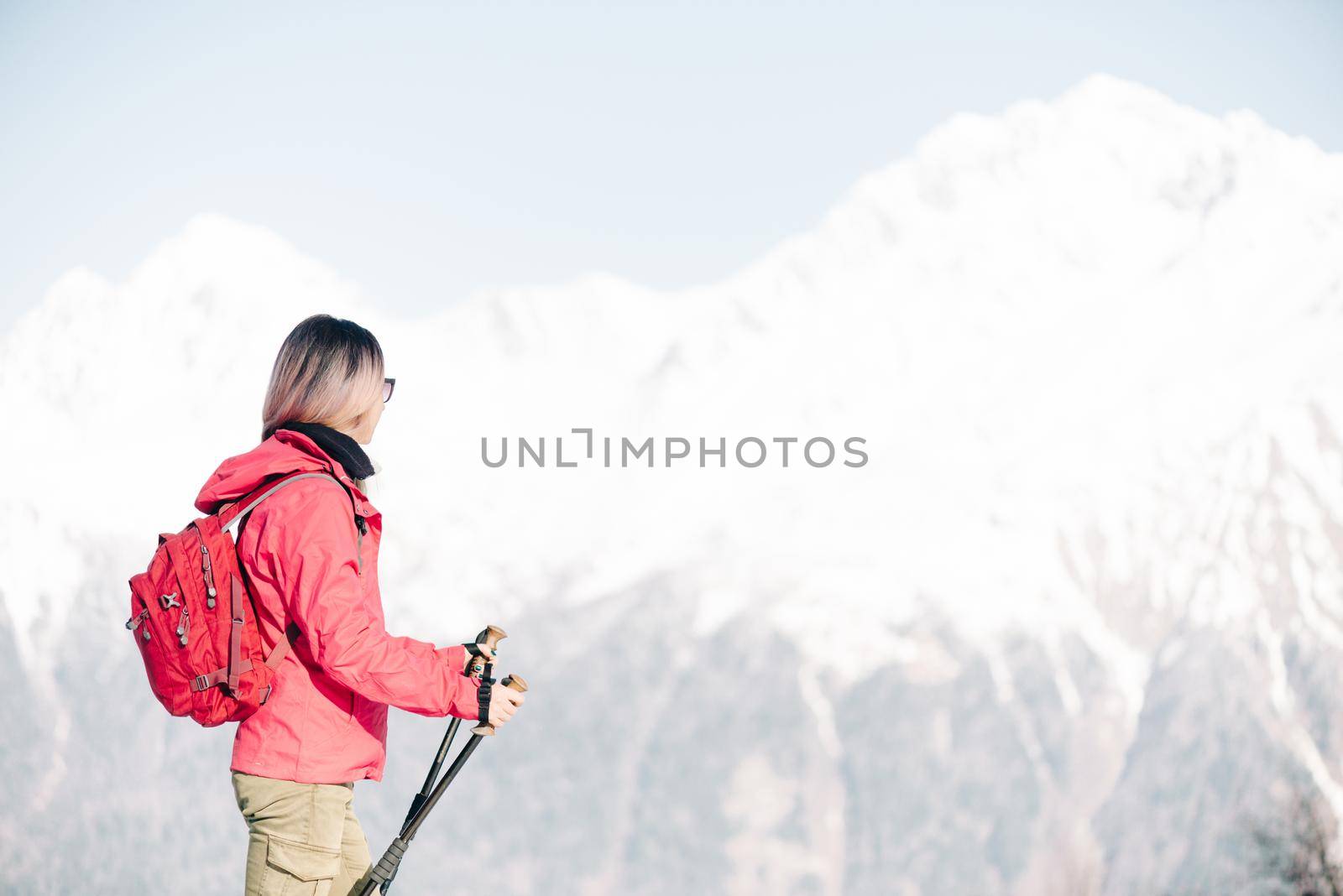 Backpacker hiker young woman with trekking poles looking at winter mountains.