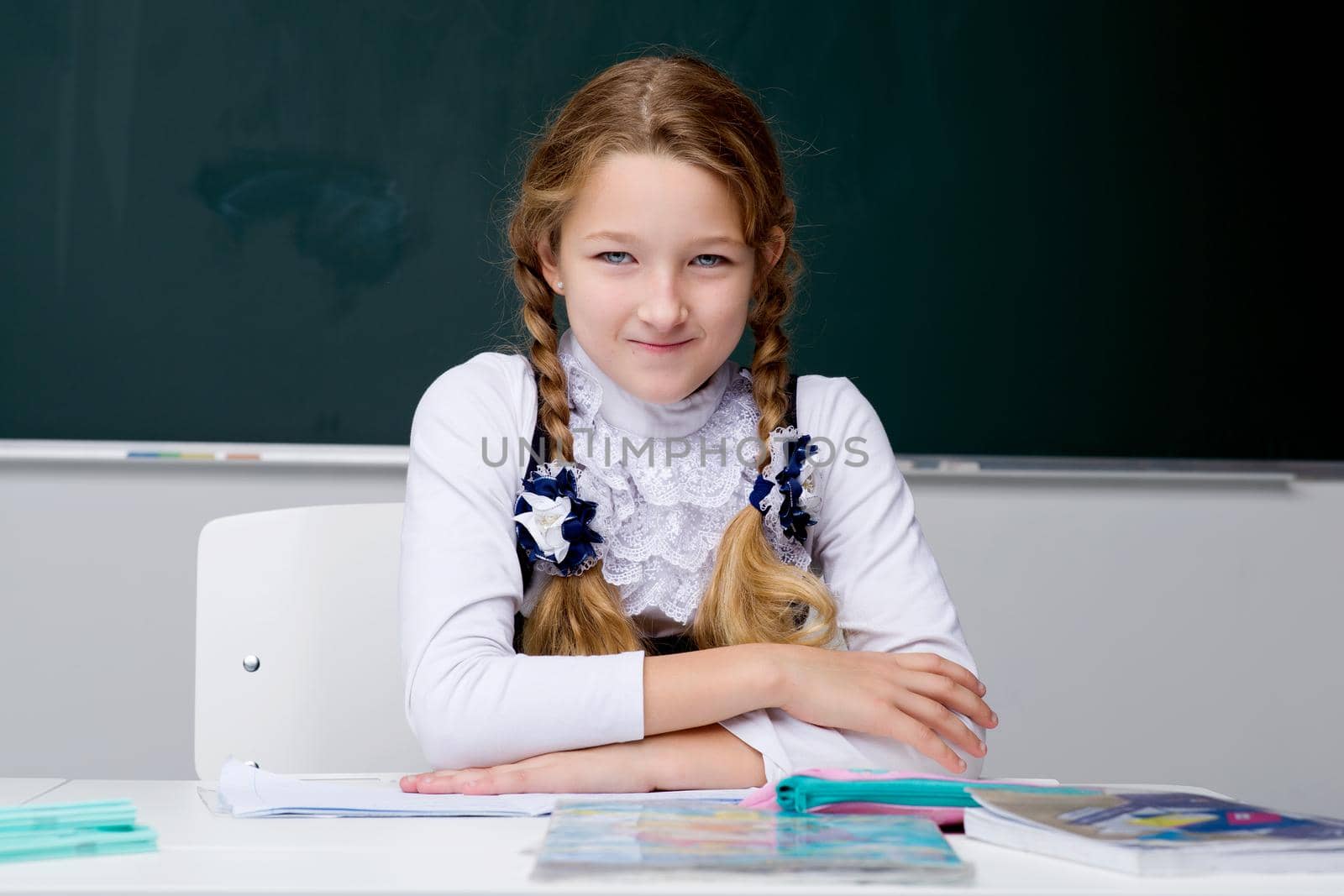 Portrait of a cute schoolgirl. A pensive, tired girl sits at a desk. Elementary school student posing in front of the blackboard. Back to school, education concept.