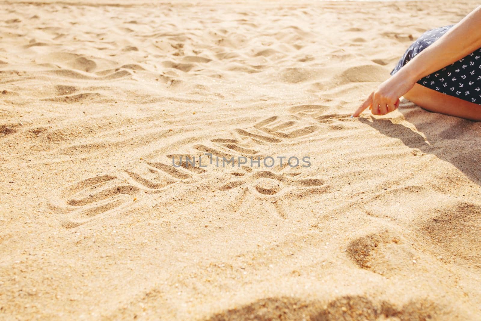 Unrecognizable young woman draws word summer on the sand with her finger, view of hand.